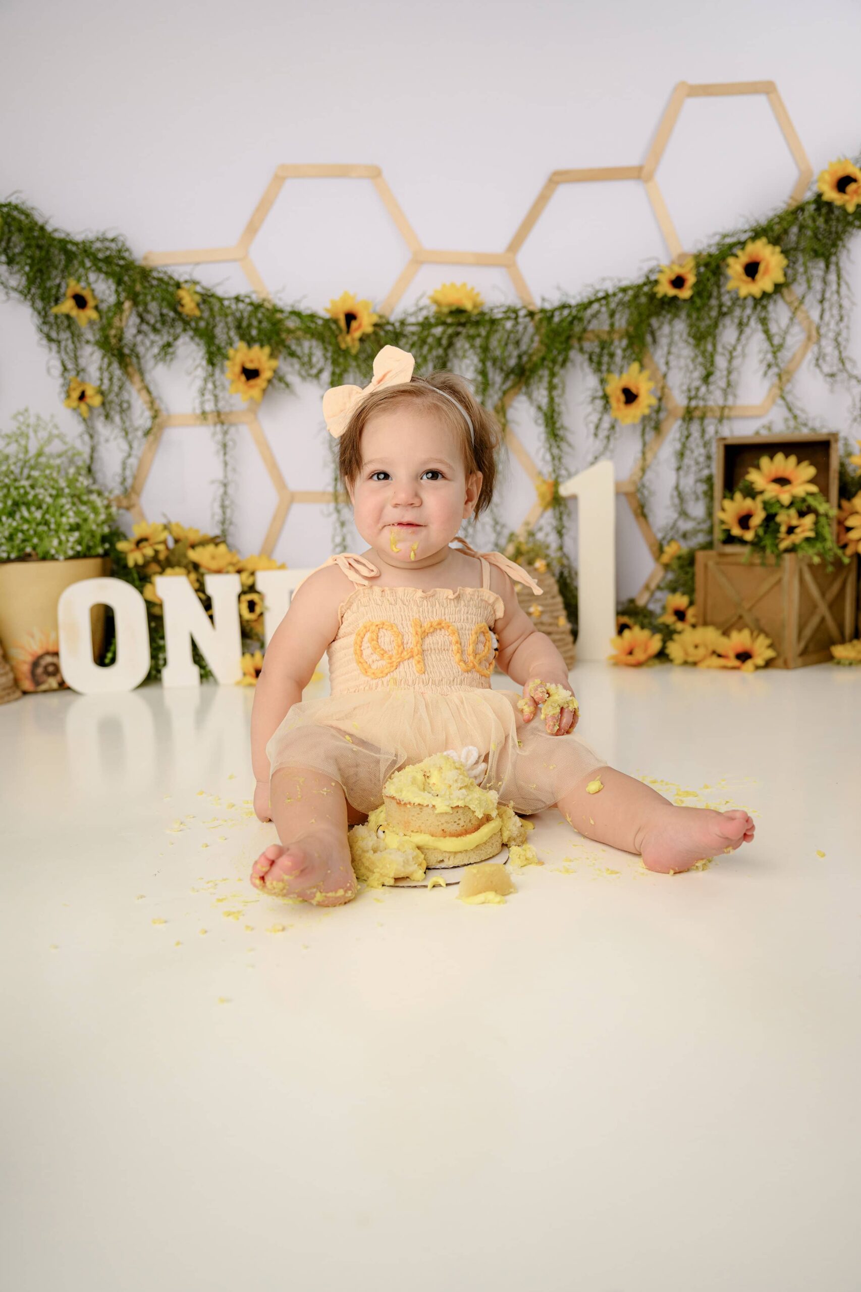 Little girl sits in her peach ONE embroidered romper looking covered in her smash cake.