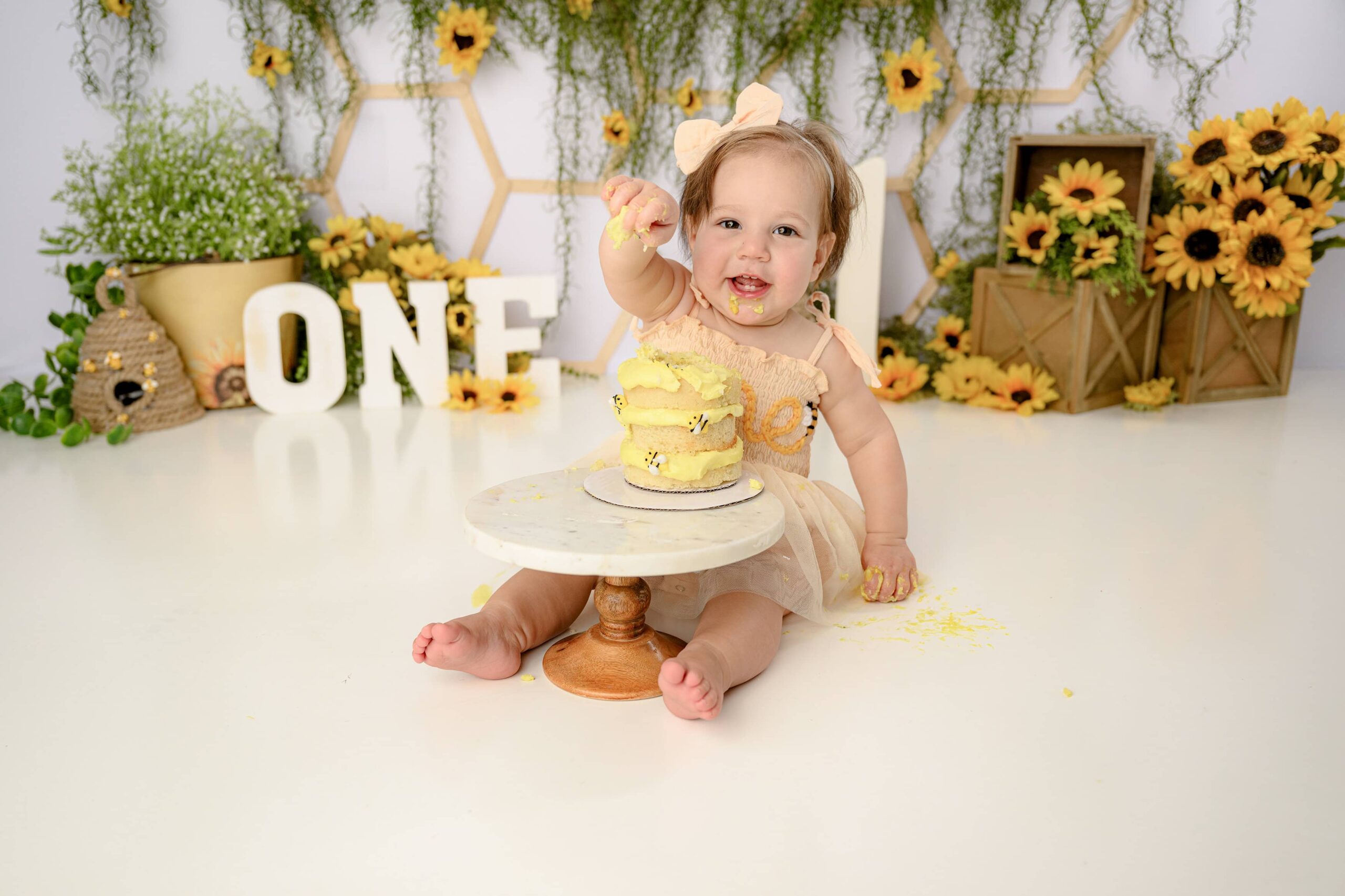 Little girl sitting with her cake on a cake stand in front of her dressed in a peach romper and a big peach bow sitting in her custom happy bee day cake smash set up