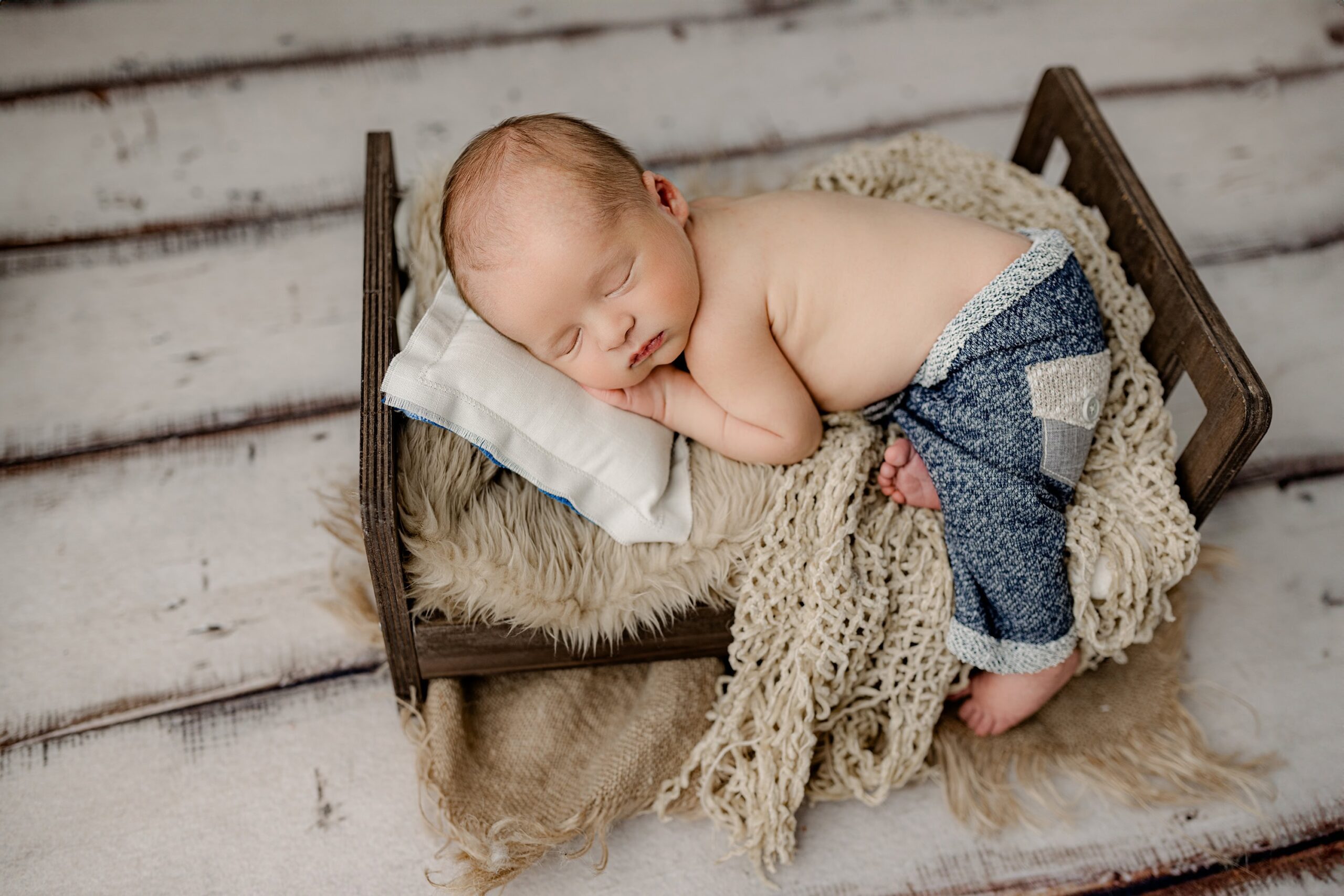 A peaceful newborn boy wearing blue pants is posed on a brown newborn bed with a cream background and floor. He is laying on his belly, one leg off the bed, with his hand resting gently under his chin, exuding calm and comfort.
