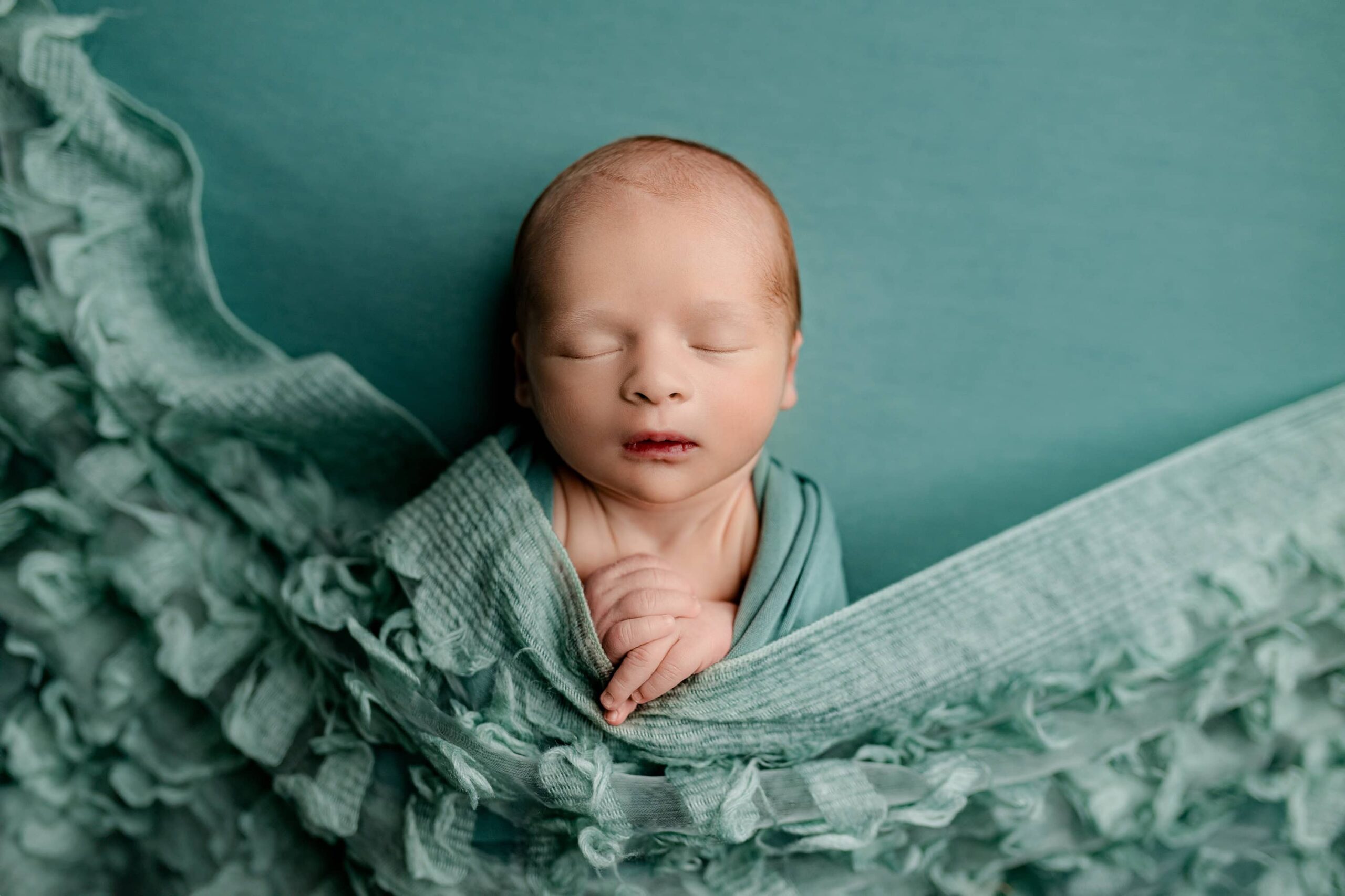 A newborn baby wrapped in an ocean teal wrap, lying peacefully on a matching teal background. The baby is gently covered with an ocean-inspired cloth that complements the serene color palette, creating a calm and soothing scene.