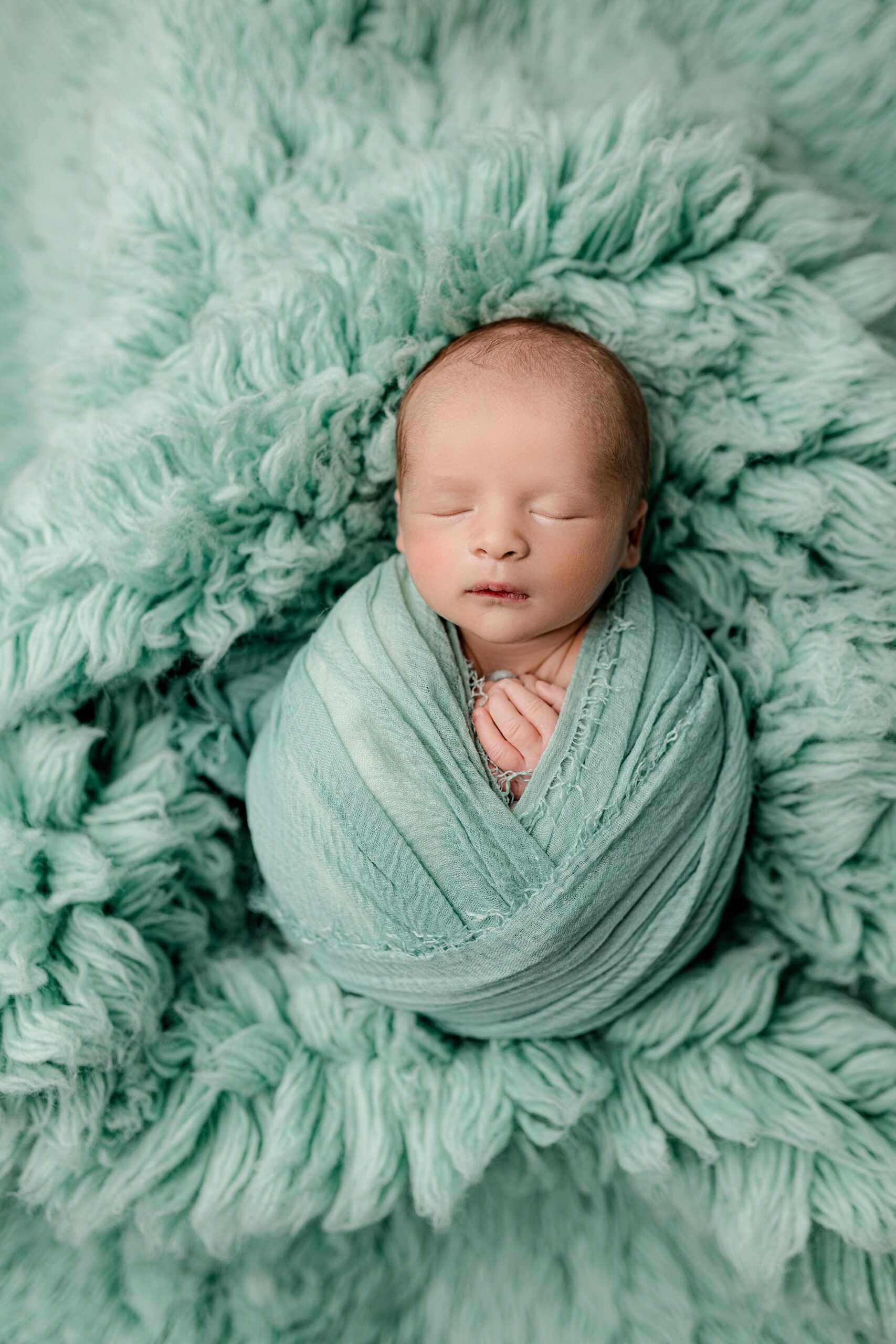 A newborn baby wrapped in a soft ocean green/mint-colored wrap, crisscrossed gently over their body. The baby’s hands are peeking out from the wrap, adding a touch of delicate detail. The serene shot is taken from above, with a matching fur background that complements the calming hues and soft textures.