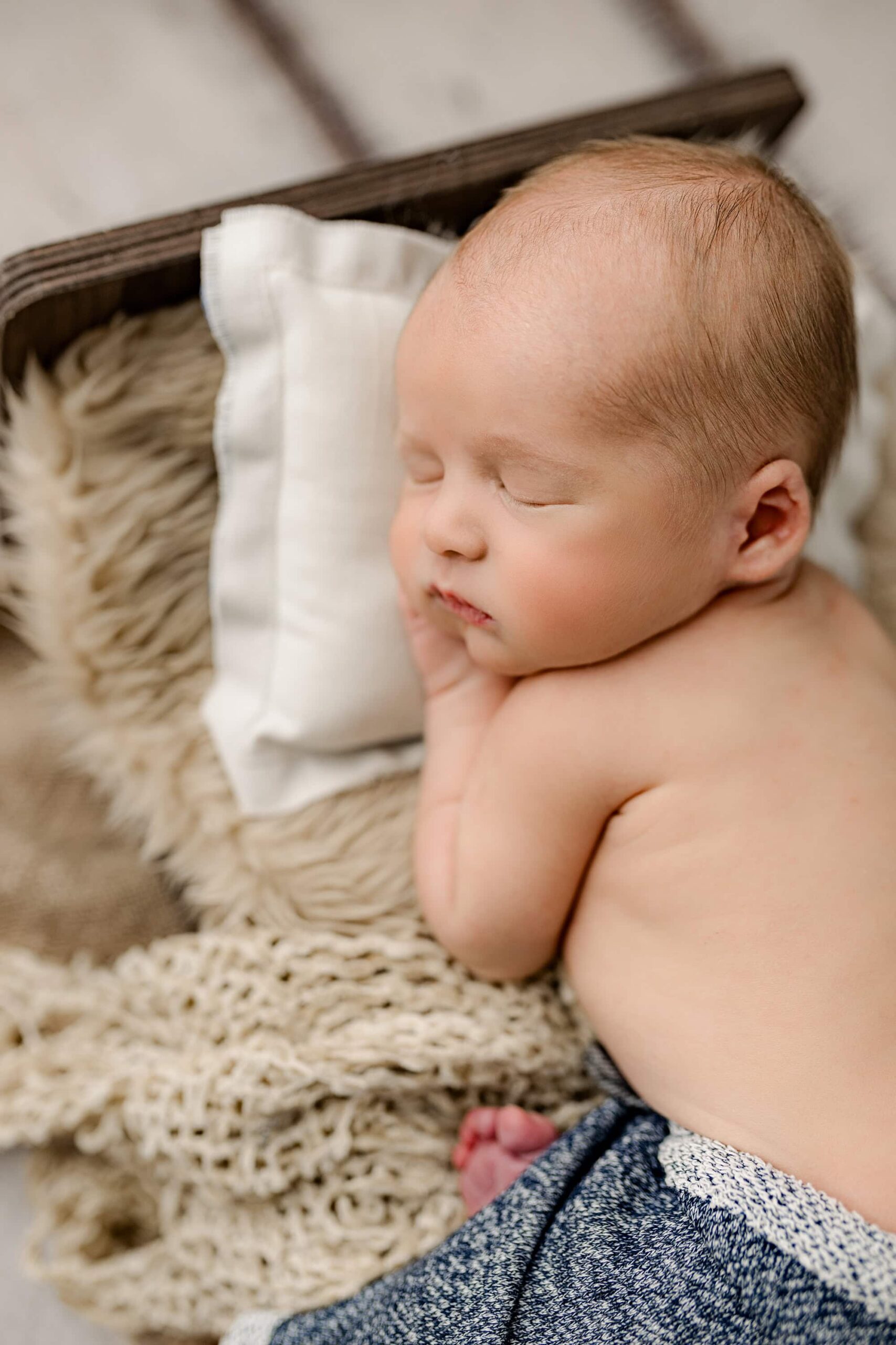 A serene overhead view of a newborn boy in blue pants, lying on his belly on a brown newborn bed. His hand is under his chin, and his profile is captured beautifully against the cream-colored background, highlighting his peaceful expression.