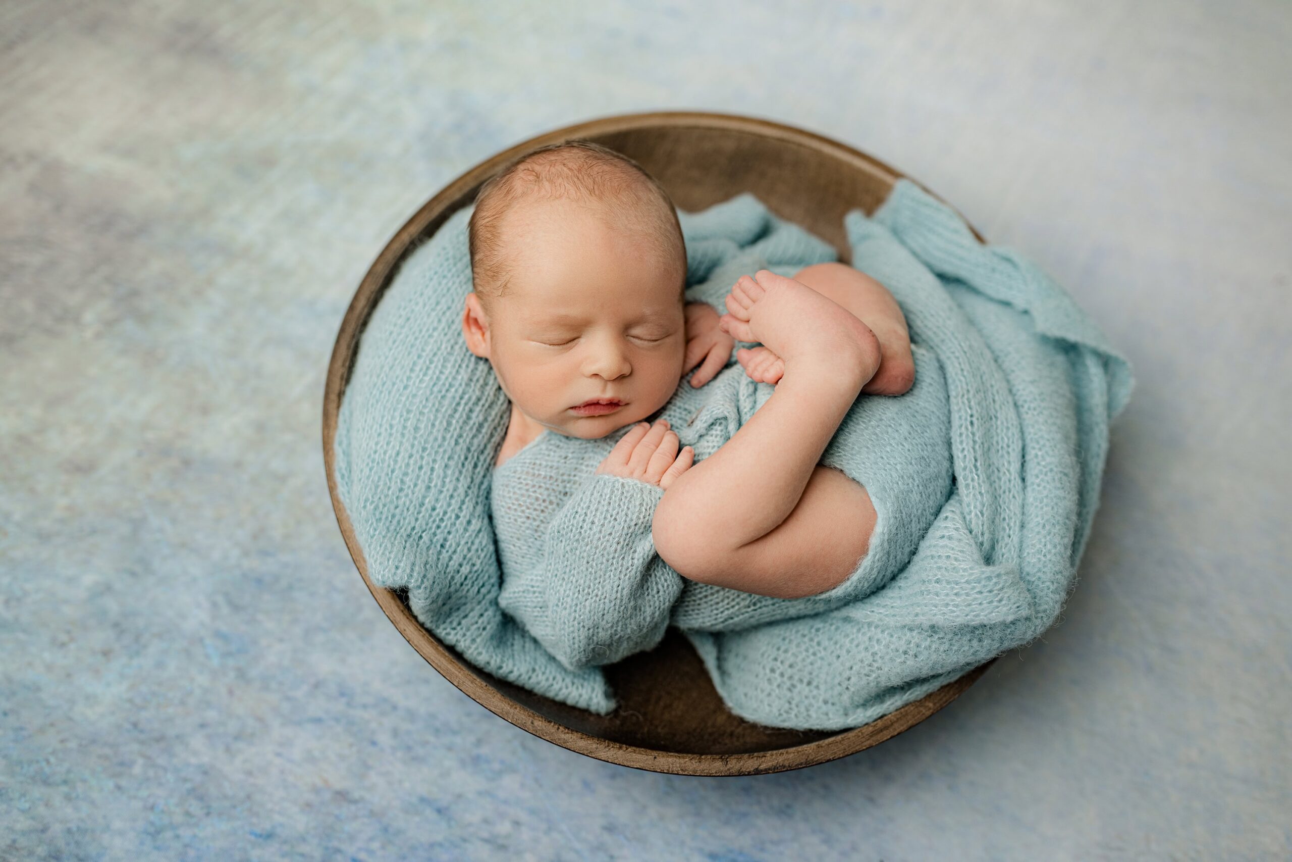 A newborn baby boy dressed in a cozy blue knit outfit, peacefully curled up in a brown bowl. The bowl rests on an ocean-inspired newborn photography drop, featuring soft shades of blue and beige. The baby is in a comfortable, snug position, exuding warmth and tranquility in this adorable pose.