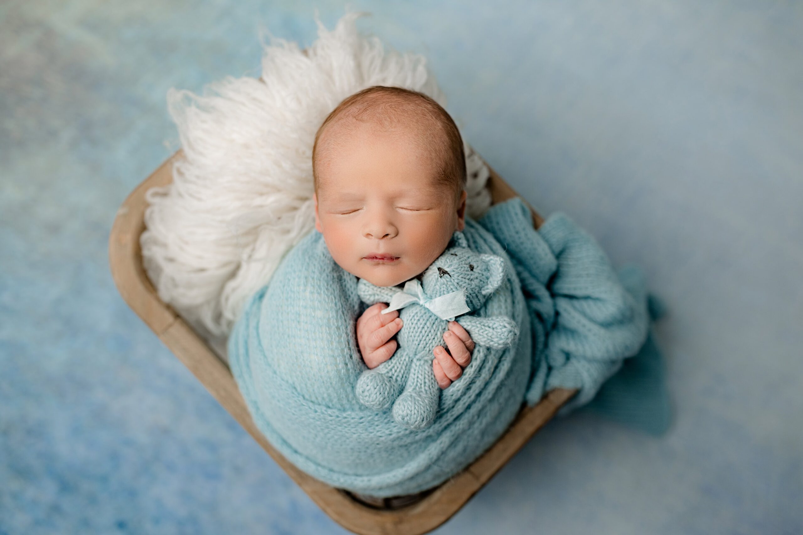 A peaceful overhead shot of a newborn baby boy wrapped in a soft mint blue wrap, nestled in a cozy basket. The background features a matching newborn photography drop in soothing shades of mint blue. The baby holds a small teddy bear, adding a sweet and sentimental touch to this calm and endearing moment.