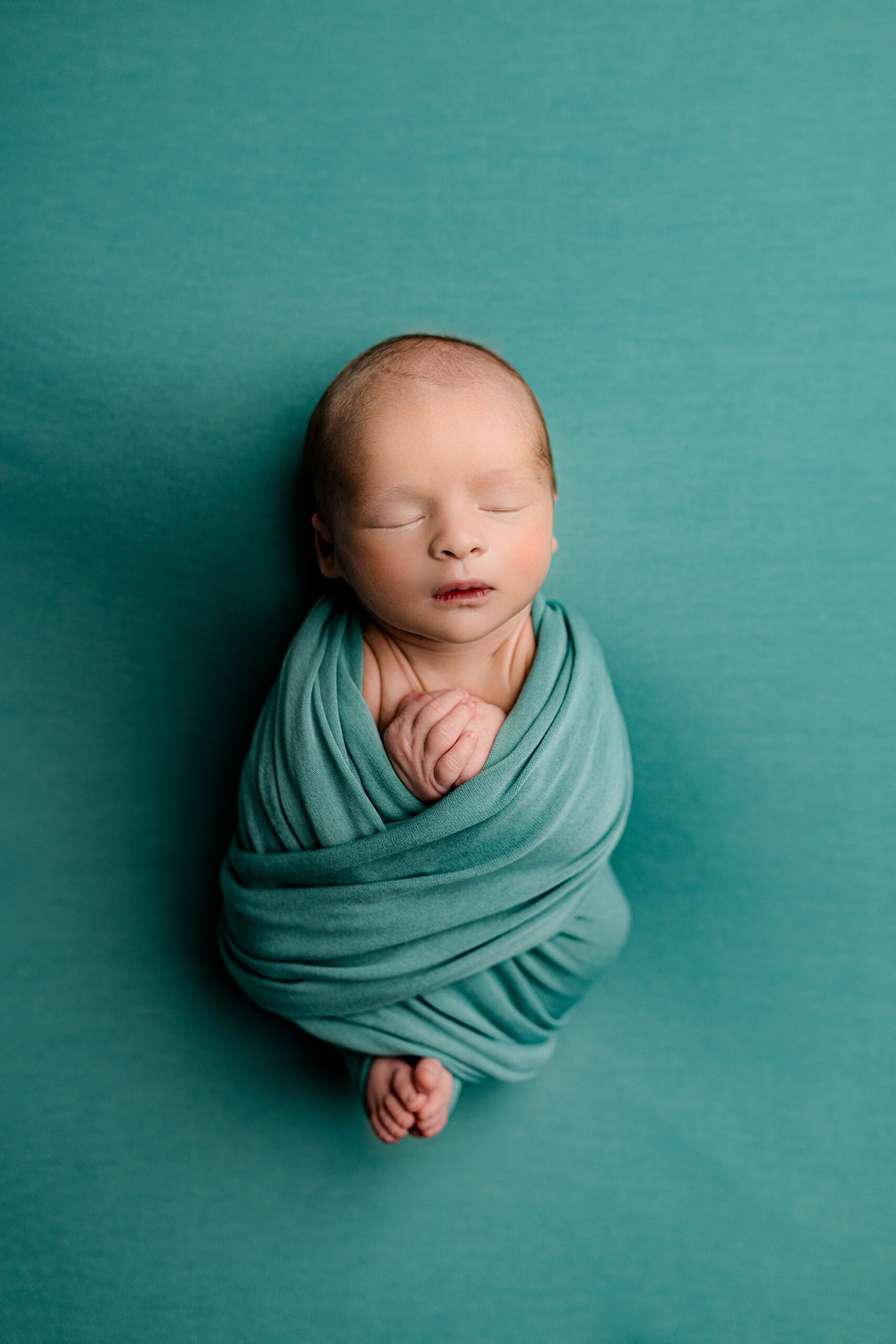 A peaceful overhead shot of a newborn baby wrapped in a soft ocean teal fabric, with the baby’s toes peeking out. The full shot showcases the baby’s serene pose, with the matching teal background enhancing the calming and harmonious atmosphere.