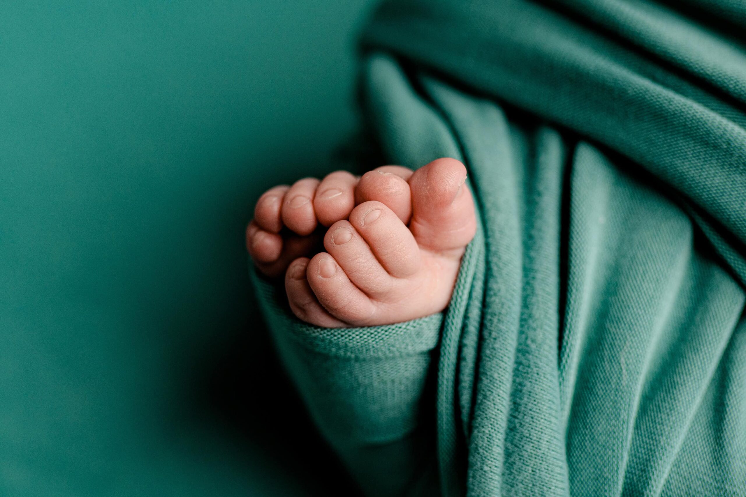 A close-up view of a newborn’s tiny toes peeking out from an ocean teal wrap. The delicate details of the toes stand out against the soft, smooth fabric, capturing the innocence and sweetness of this precious moment.