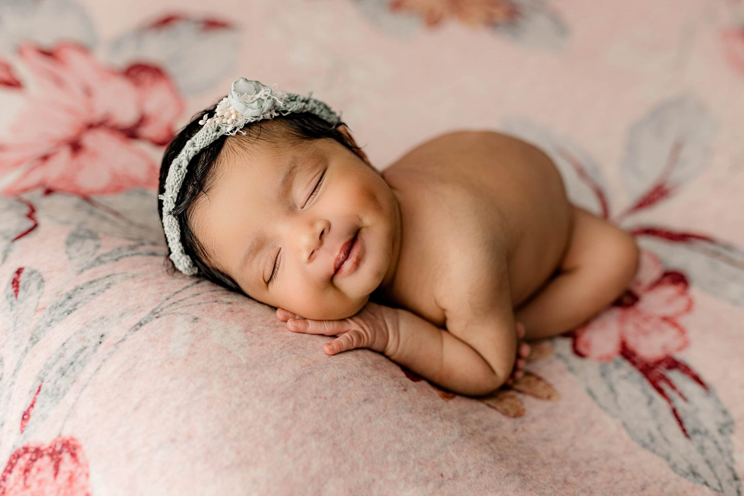 A newborn baby girl with a head full of soft black hair is posed on a floral background in shades of teal and pink. She is curled into the "taco position," with her face forward and her body gently tucked. She wears a delicate teal headband, accentuating her peaceful expression.