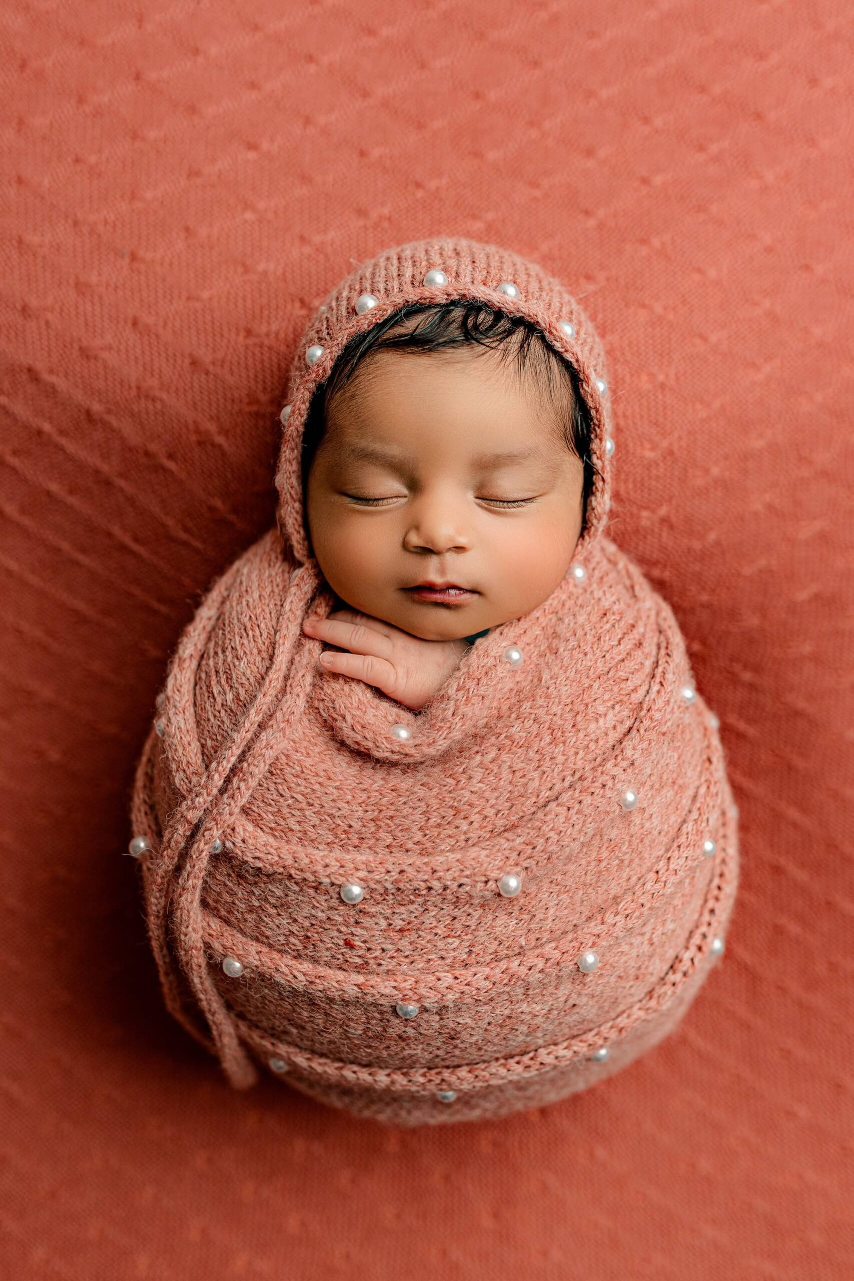 The newborn baby girl is wrapped in a delicate salmon-colored wrap adorned with tiny pearls, lying on her back in a soft, round shape. She wears a matching bonnet embellished with pearls, perfectly complementing the monochromatic salmon background. Her peaceful expression and the elegant details create a timeless and dreamy image.
