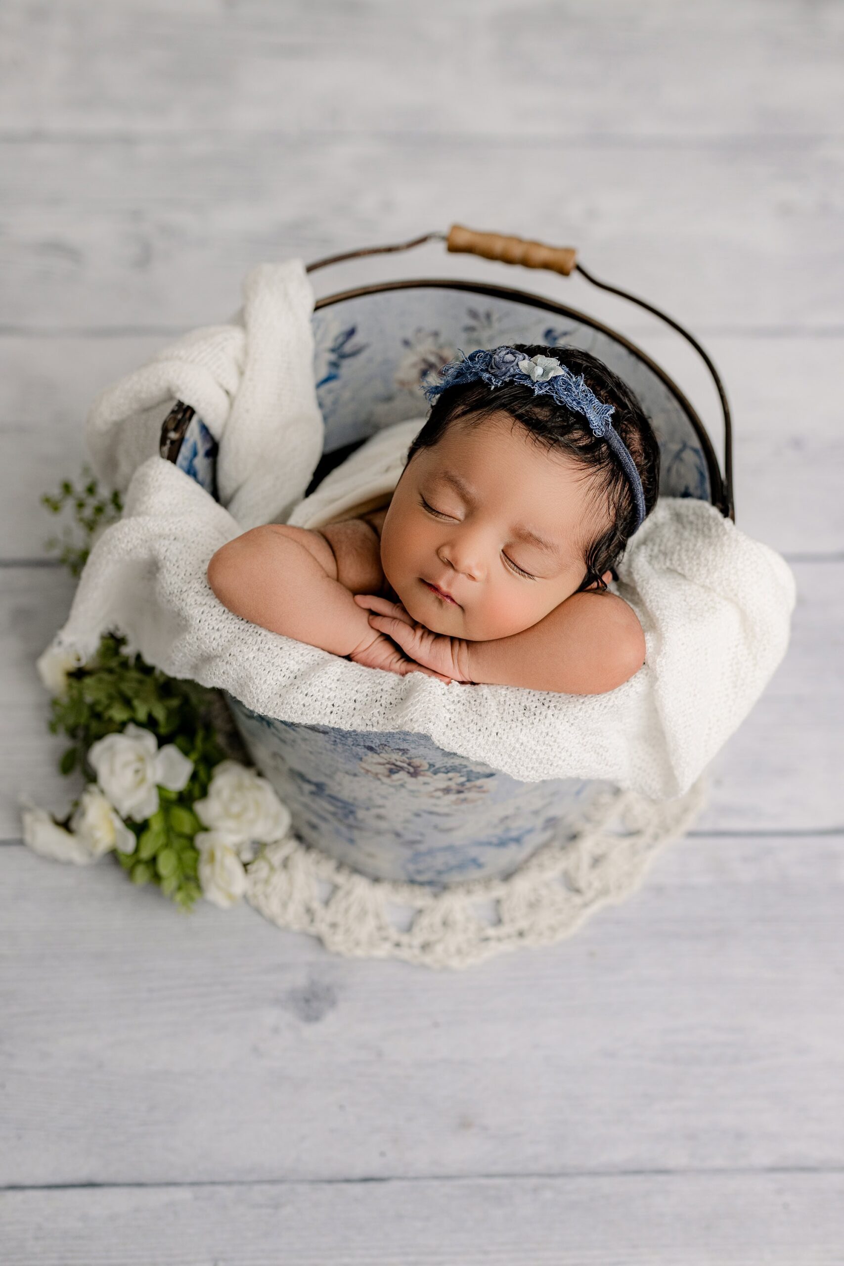 The baby girl is nestled in the same blue floral bucket, with her hands gently tucked under her chin on a white wrap. Her blue headband highlights her delicate features, while the portrait orientation draws focus to her sweet expression and the intimate details of the setup. The grey and white flooring adds a subtle, timeless backdrop to the scene.