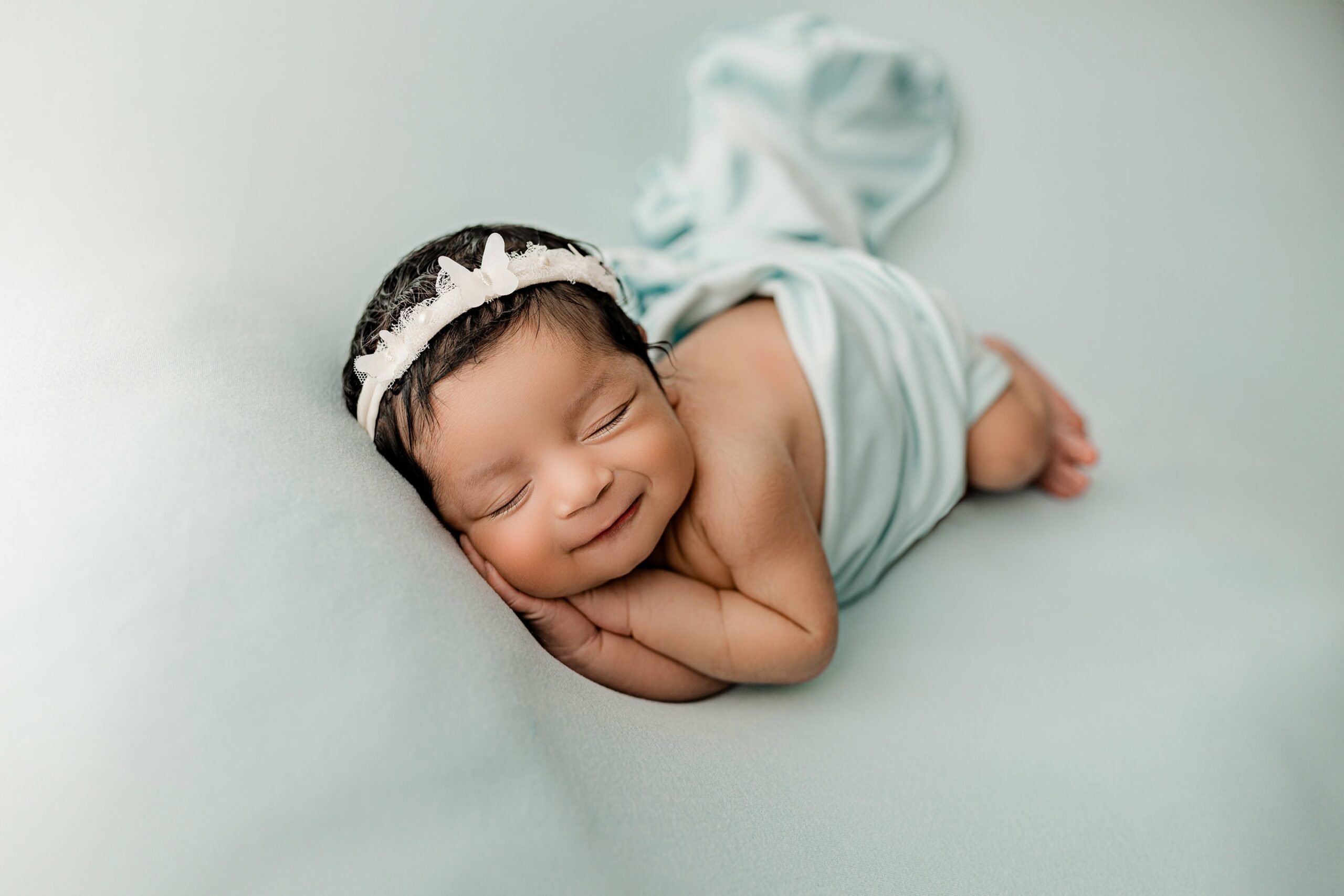 The newborn baby girl is posed on a soft mint blue background, lying on her right side with her tiny hands gently tucked under her cheek. She wears a delicate butterfly headband that adds a whimsical and graceful touch to the serene setup. Her peaceful expression and the pastel tones create a dreamy, soothing portrait.