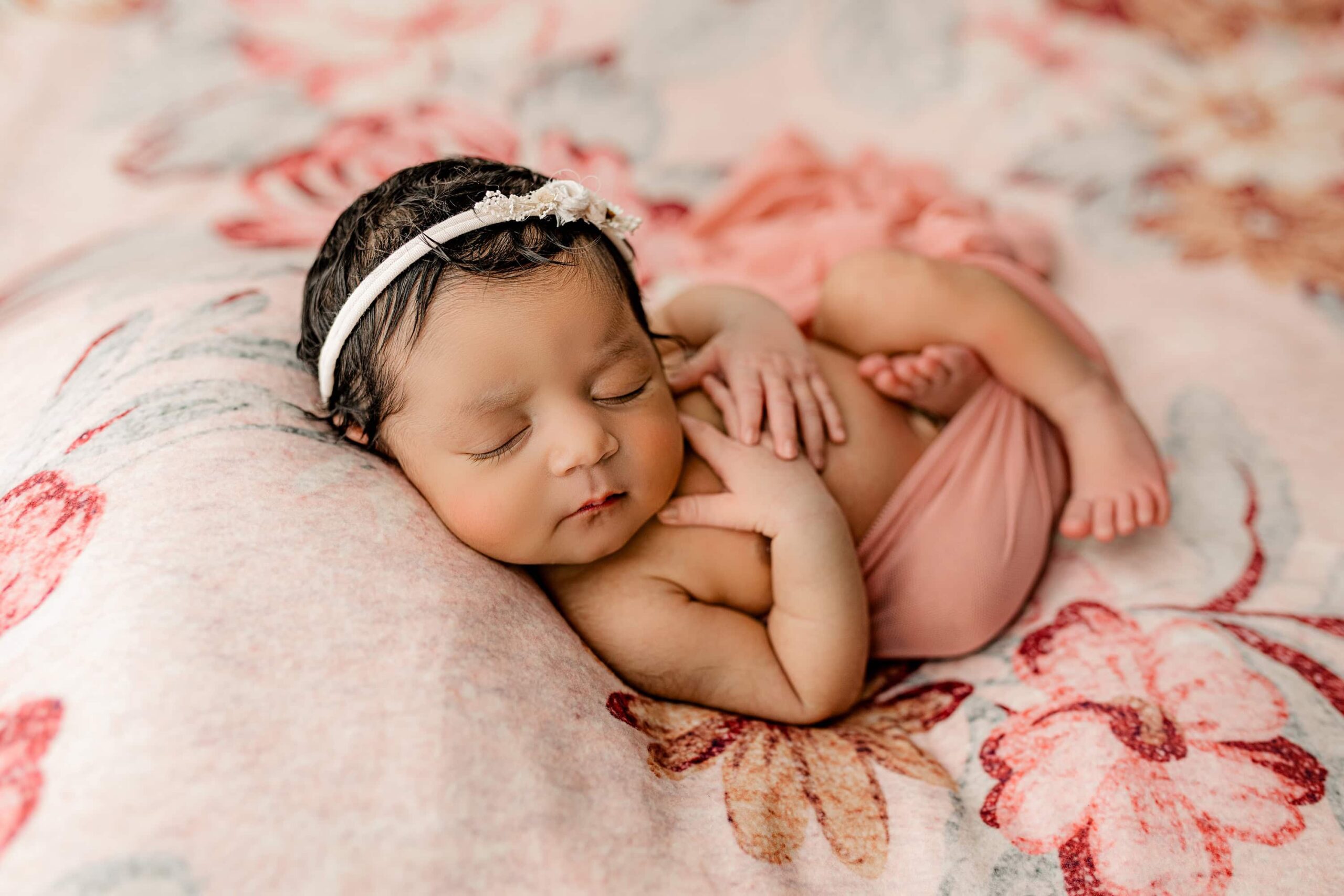 The baby is now posed on her back, gently wrapped in a sheer pink fabric that showcases her tiny frame. Her whole body is visible, and she wears a cream headband that adds a touch of elegance to the setup. The teal and pink floral backdrop enhances the dreamy composition and compliments her skinton.