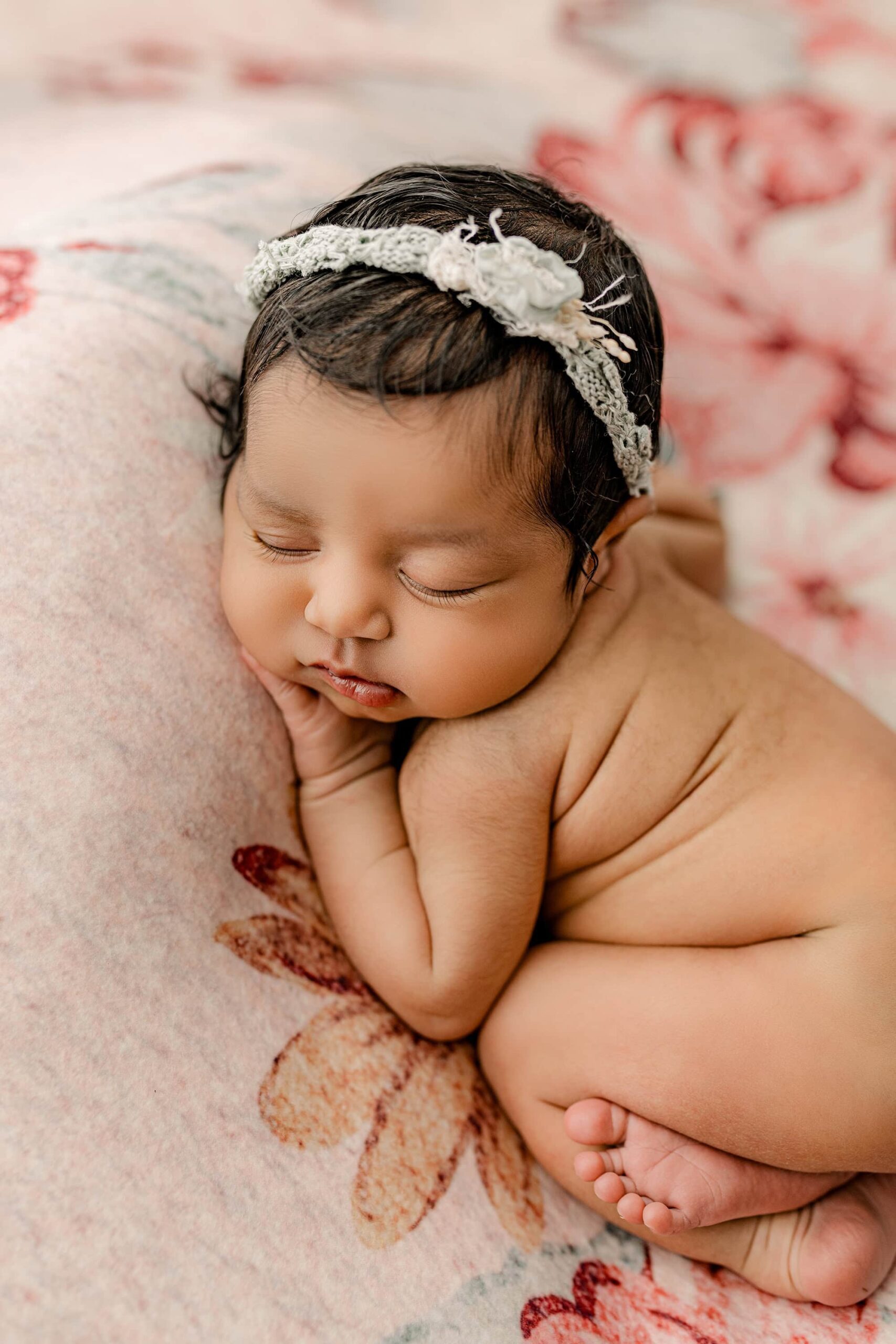 Another overhead image of the baby girl, this time posed with her knees curled under her. Her little chubby rolls are beautifully highlighted, and she wears a matching teal headband that complements the floral backdrop.