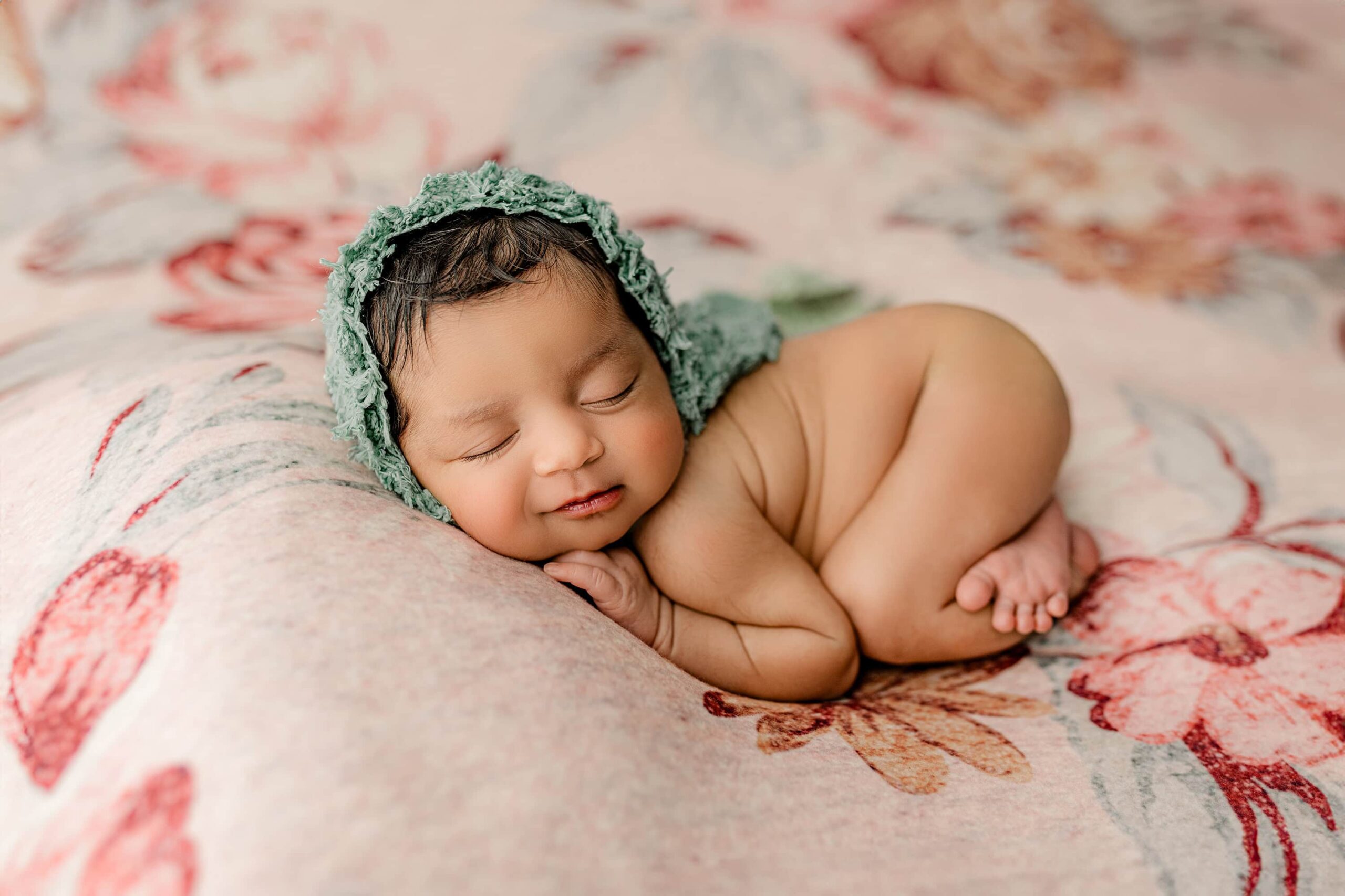 The baby is now posed on her belly with her knees tucked under her and her bum in the air. She’s wearing a teal bonnet and flashing a subtle smile. Her entire body is visible, and her peaceful demeanor radiates warmth and charm.