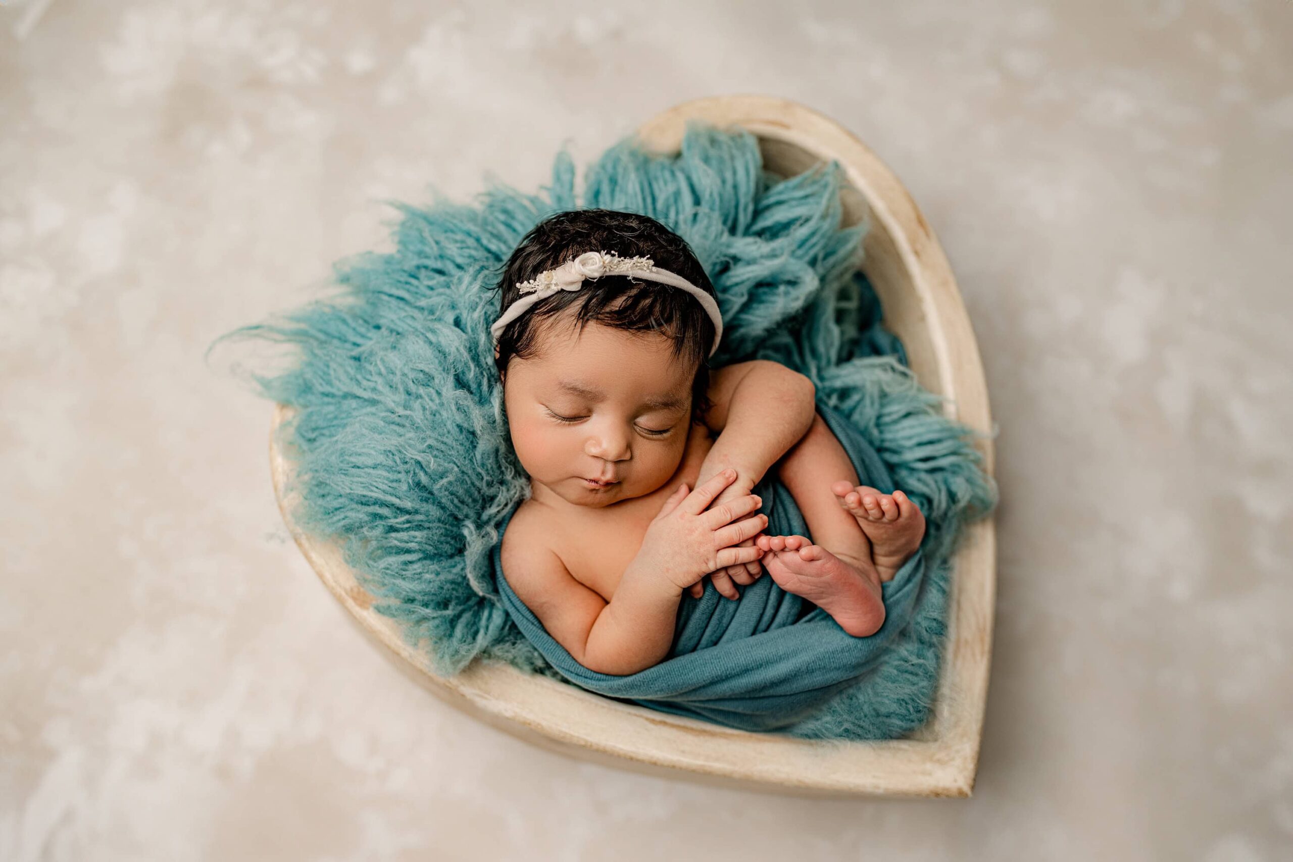 The baby girl purses her lips as she lays in a cream heart-shaped bowl, swaddled in a teal wrap that highlights her tiny frame. Her head is slightly tilted, framed by her thick black hair and a coordinating teal headband. The simplicity of the cream painting like background setup draws attention to her sweet, serene expression.