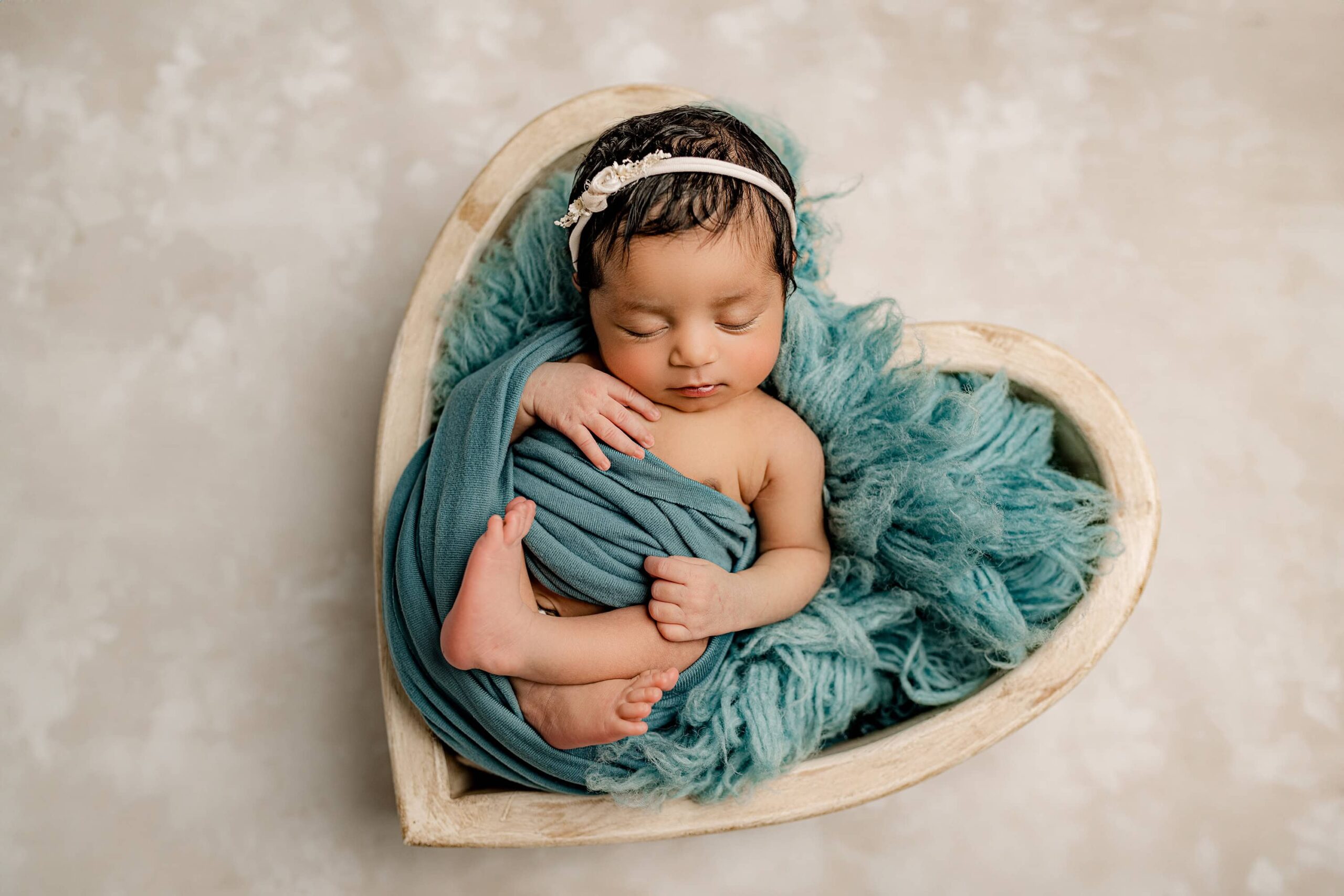 Newborn photo of a baby girl nestled in a cream-colored heart-shaped bowl, wrapped snuggly in a soft teal fabric. Her head rests gently on the wrap, showcasing her delicate features and thick black hair. She wears a matching teal headband, and the setup exudes a serene and cozy vibe.