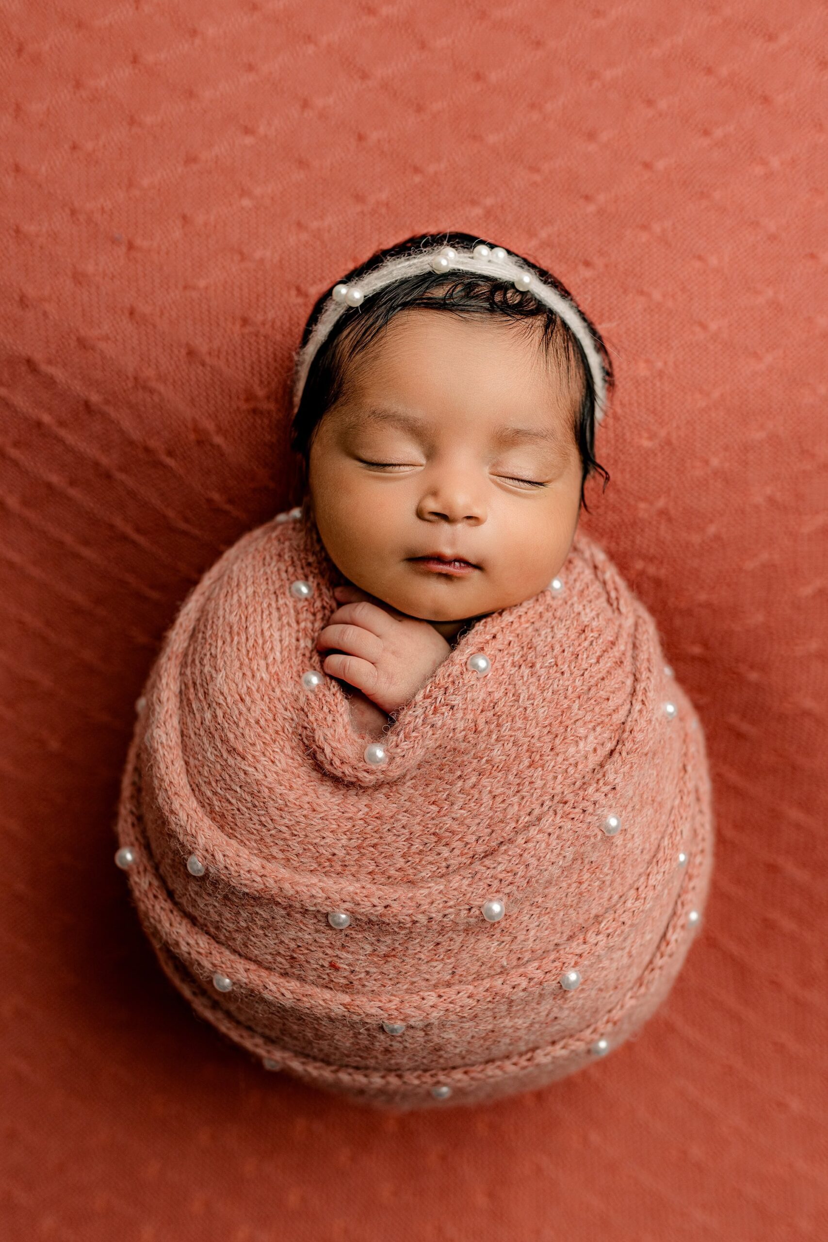 The baby girl is posed on the same salmon-colored background, wrapped snugly in the pearl-accented wrap. This time, she wears a cream headband that adds a subtle contrast to the warm tones. Her tiny frame and serene pose are highlighted beautifully against the cohesive color palette.