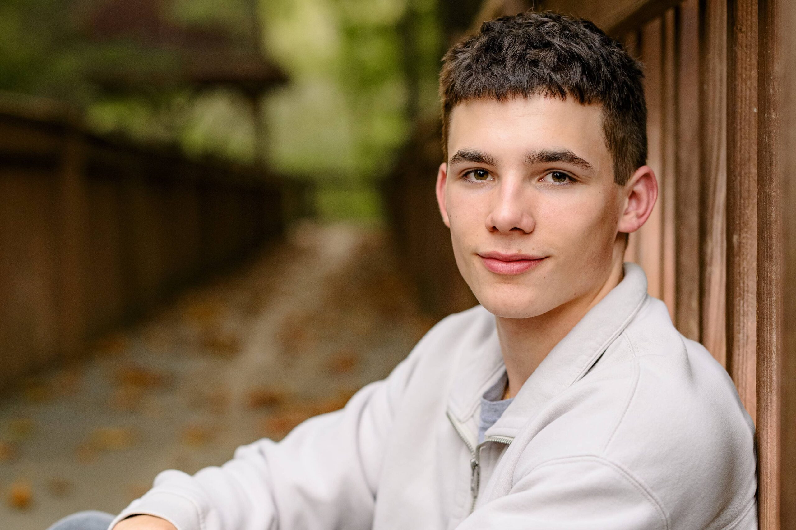 senior boy stands with his hands in his pockets wearing a cream crew neck shirt and grey pants sitting with his arms down