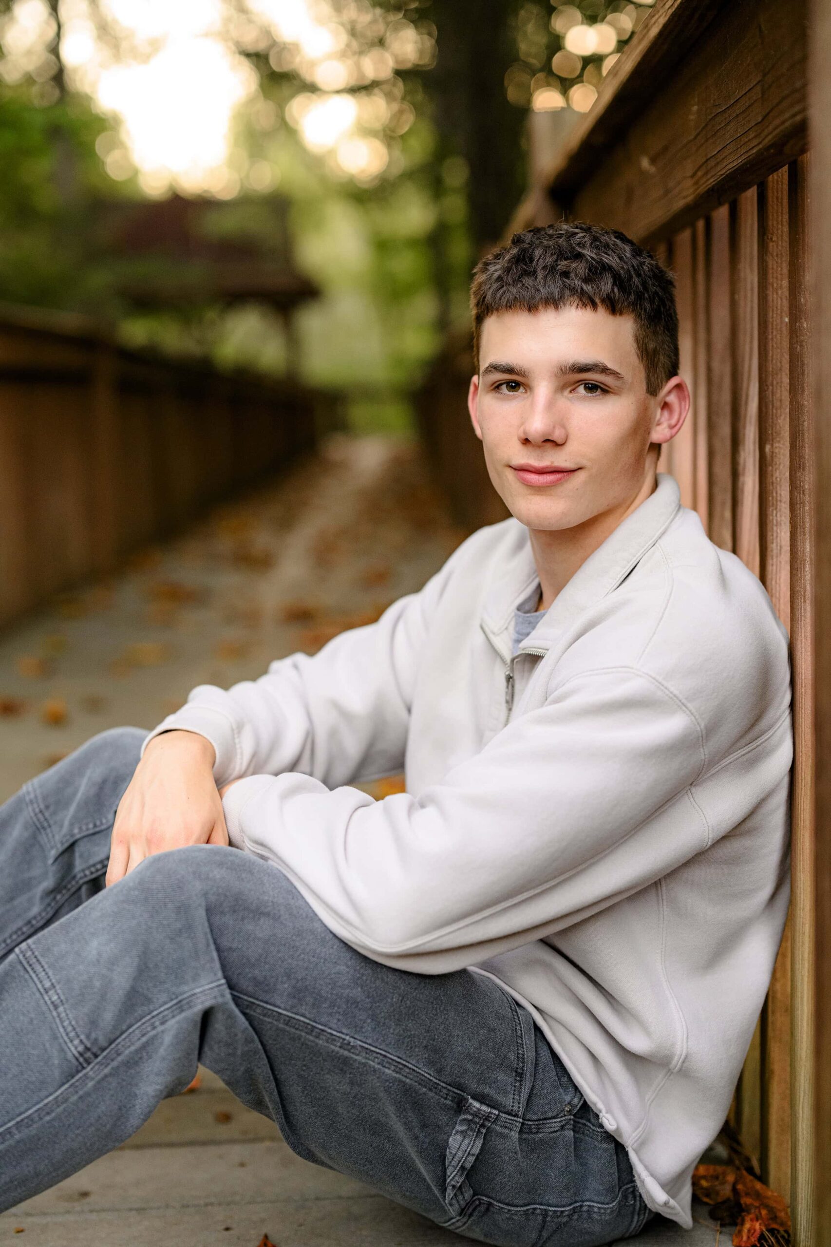 senior boy stands with his hands in his pockets wearing a cream crew neck shirt and grey pants sitting with his arms down