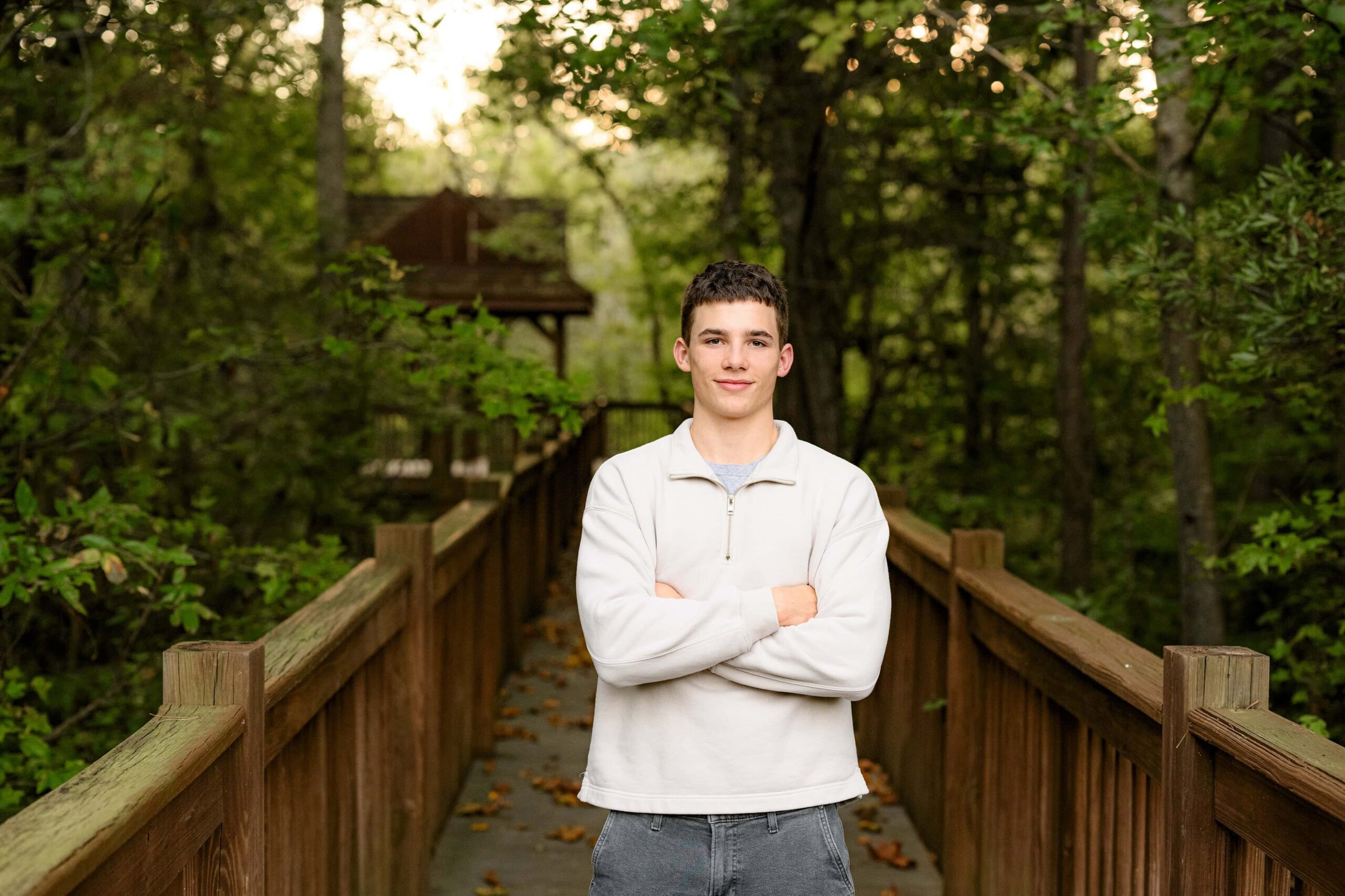 senior boy stands with his hands in his pockets wearing a cream crew neck shirt and grey pants standing with his arms crossed