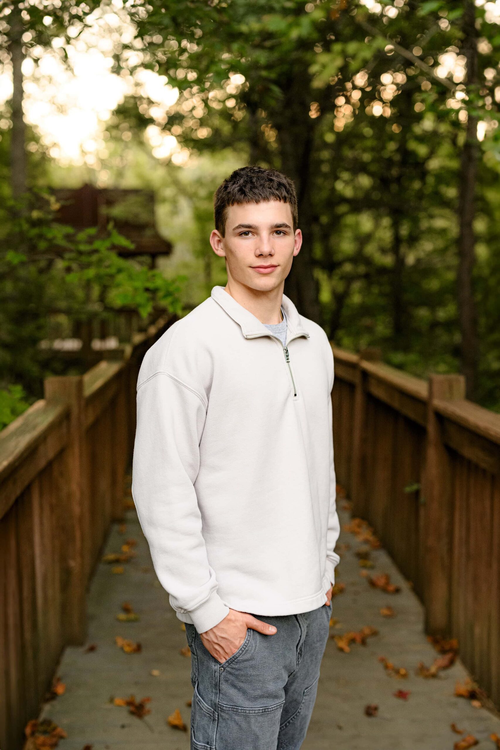 senior boy stands with his hands in his pockets wearing a cream crew neck shirt and grey pants on a walkway