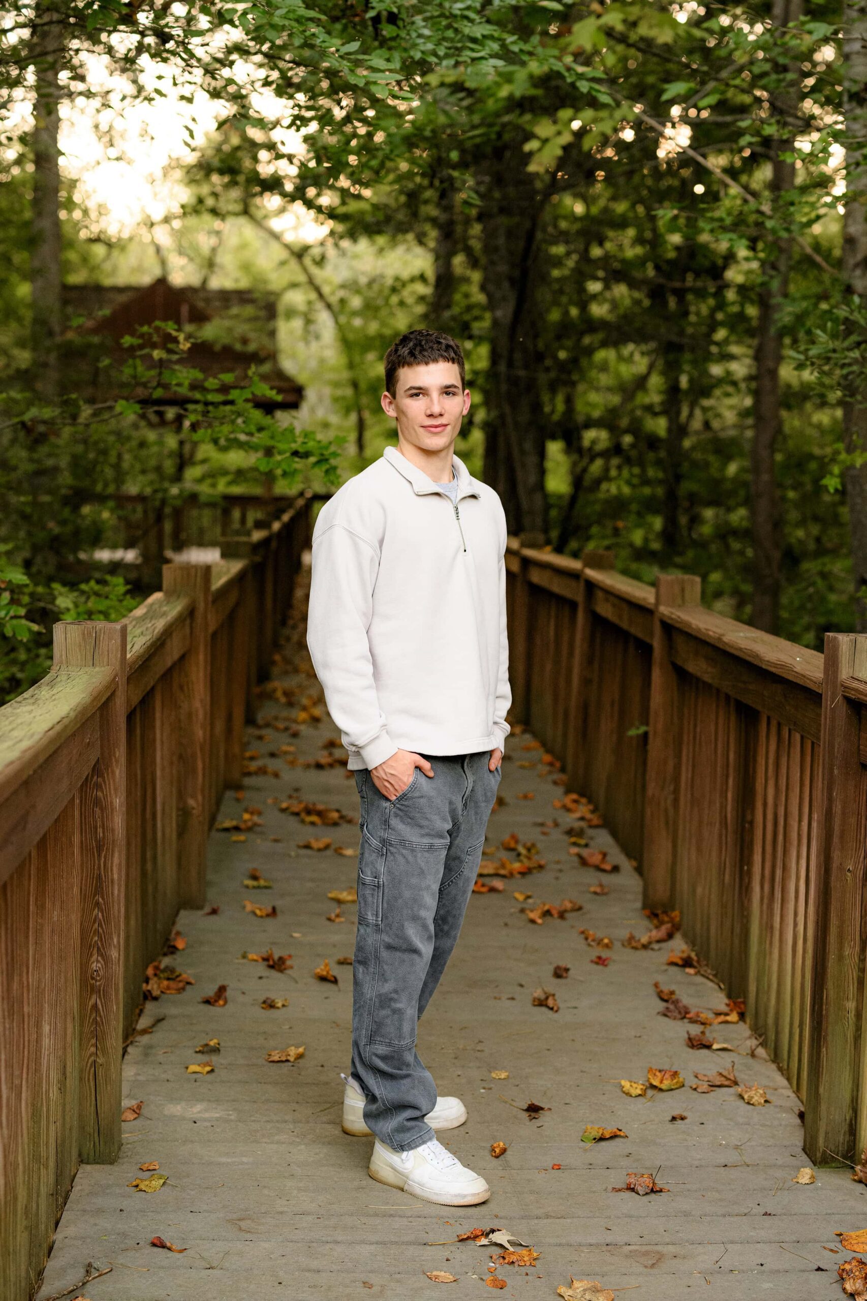 senior boy stands with his hands in his pockets wearing a cream crew neck shirt and grey pants
