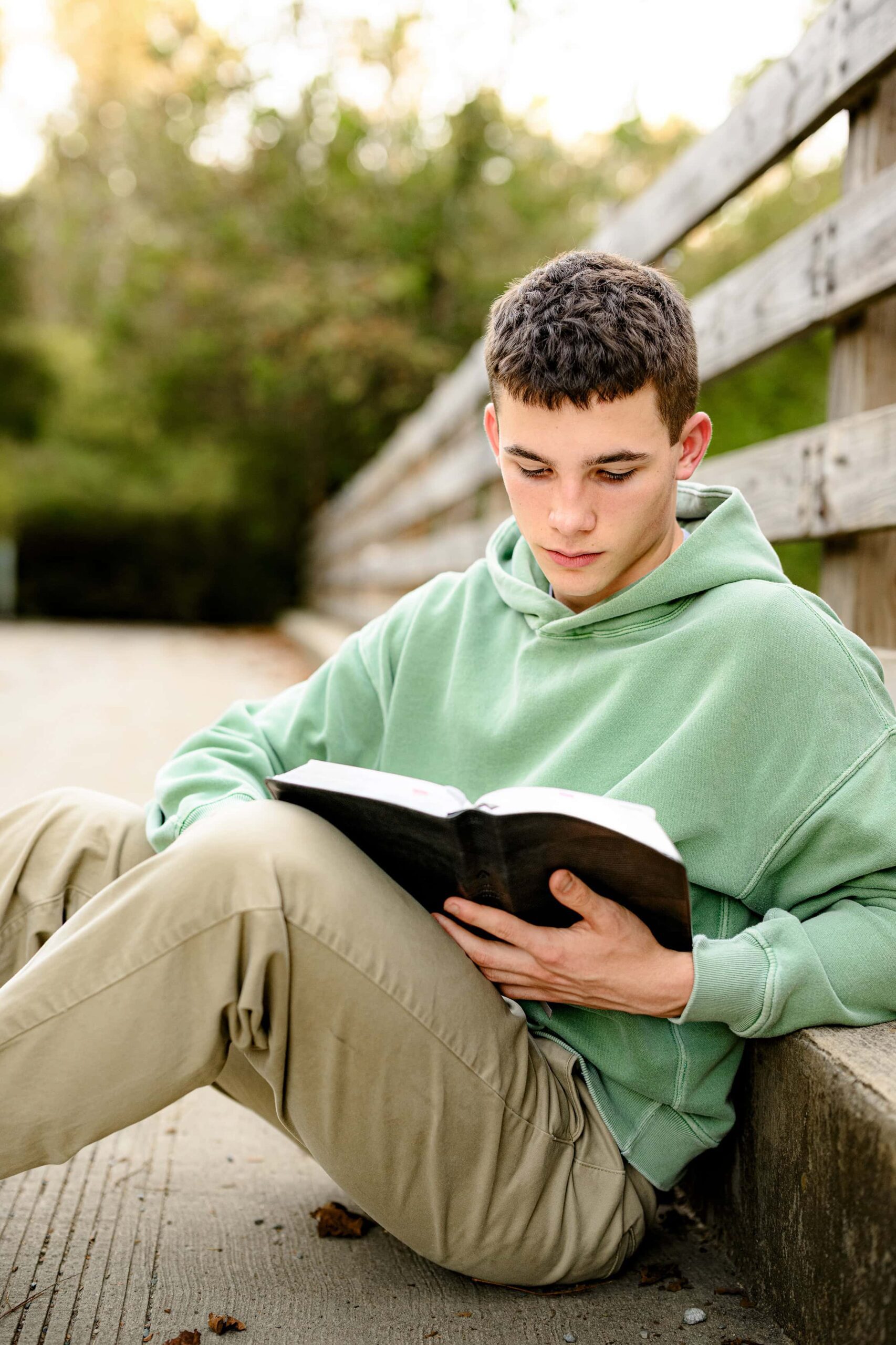 senior boy sits reading his bible while wearing a green shirt and brown pants with a meaningful senior session prop