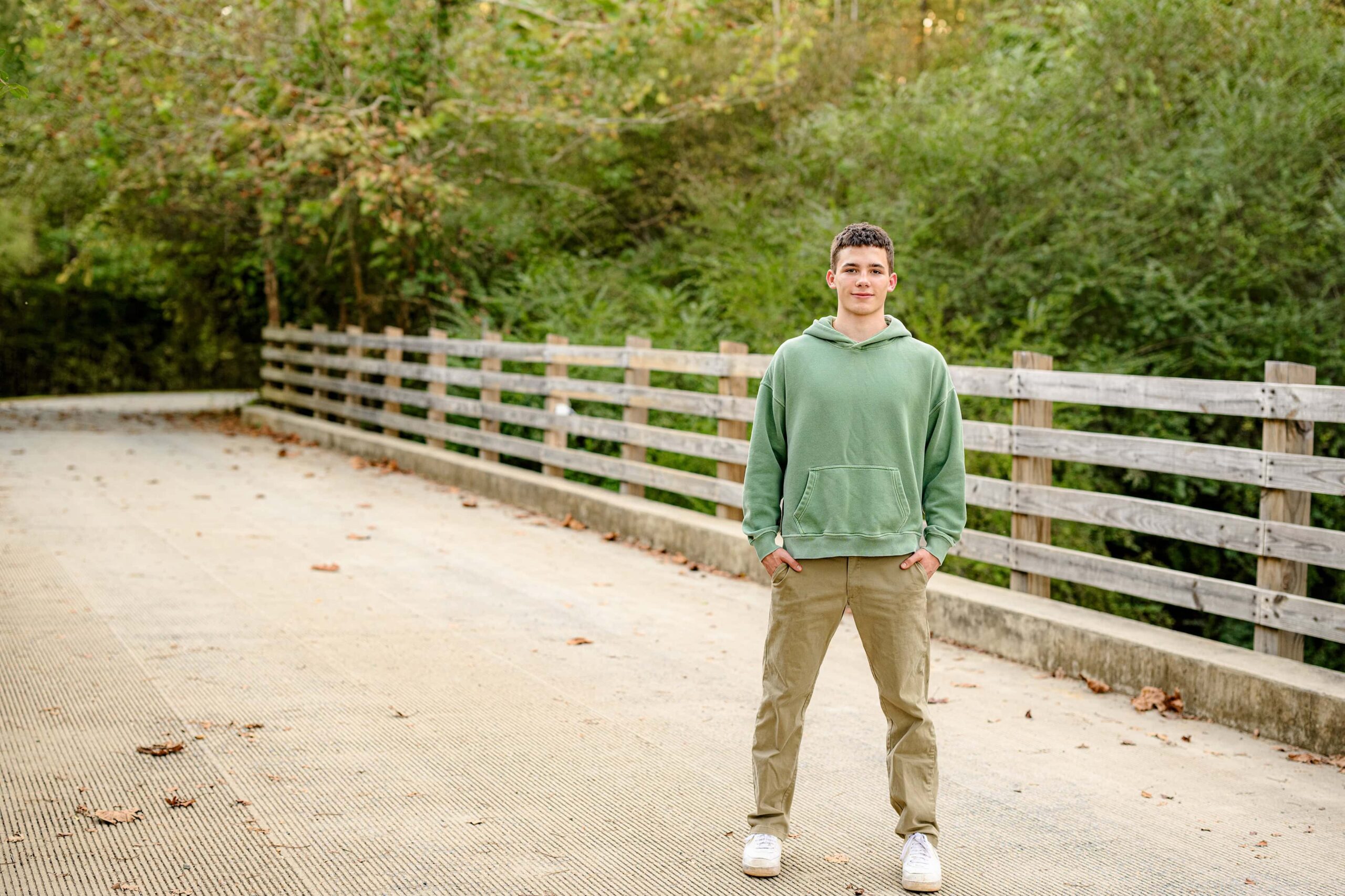 senior boy stands on bridge wearing a green shirt and brown pants.