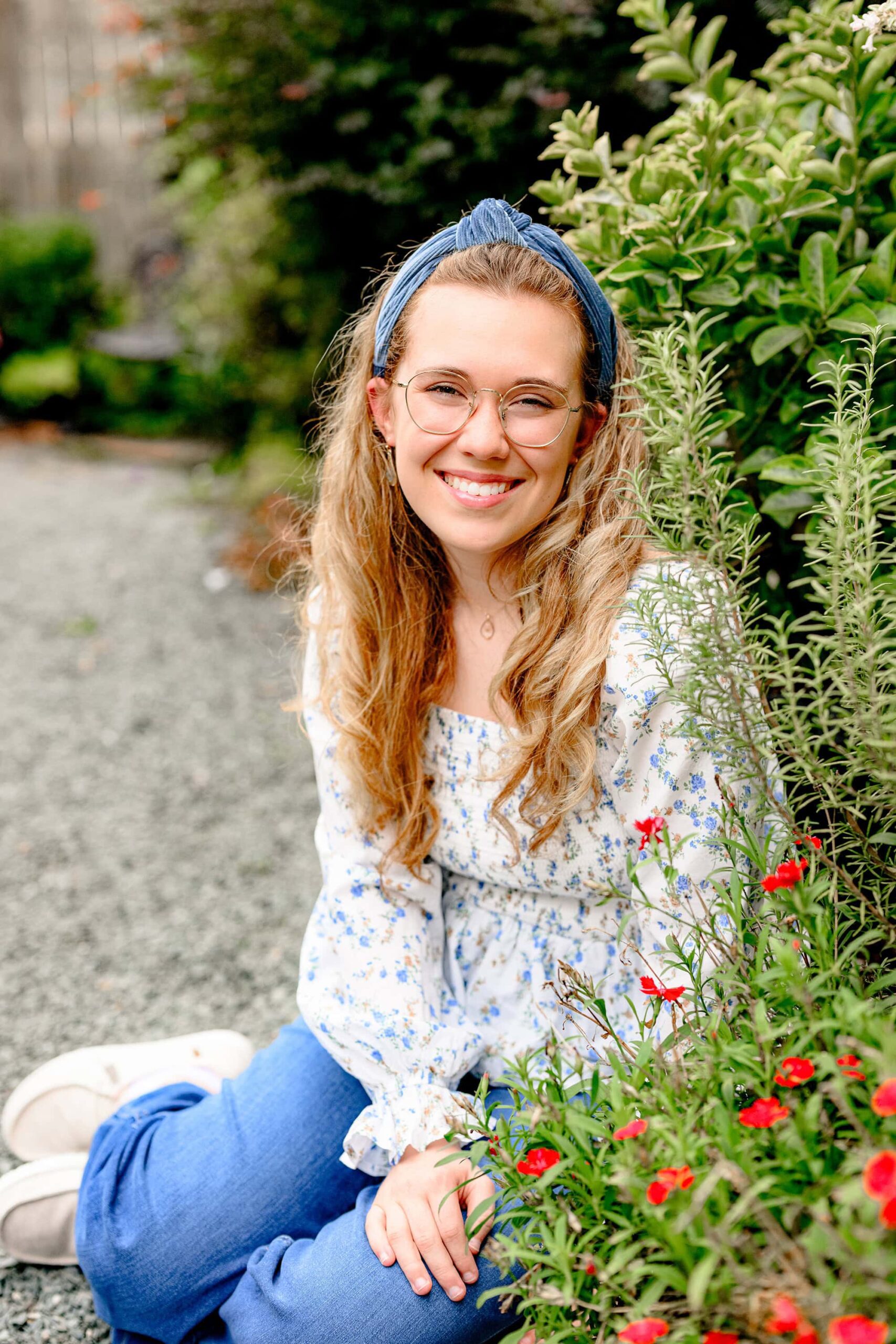 girl sits in a flower garden dressed in jeans and a flower top wearing her glasses and smiling