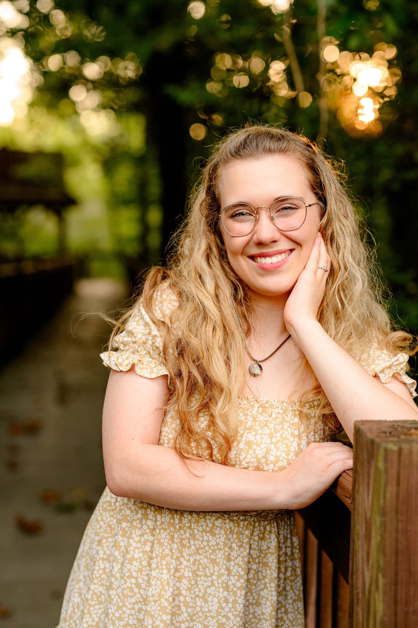 senior girl poses on a on wooden walkway wearing a brown dress and shoes with her hand by her face as the sun sits behind her making her hair glow
