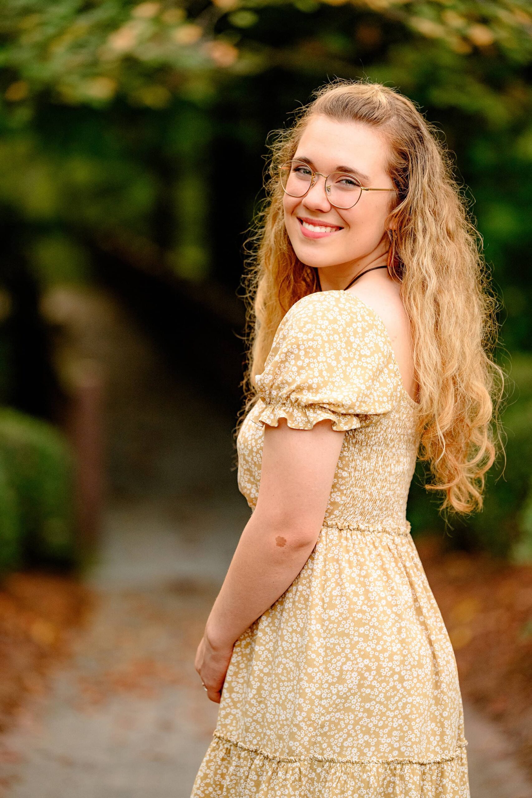 looks over her shoulder while standing close to a walkway wearing a brown dress during her senior portrait session in waxhaw, nc.