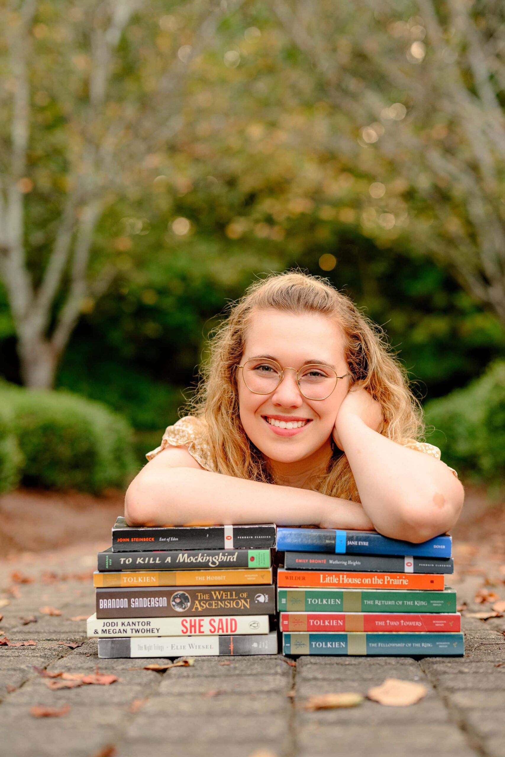 girl leaning on a stack of her favorite book wearing her glasses and smiling during her senior portrait session