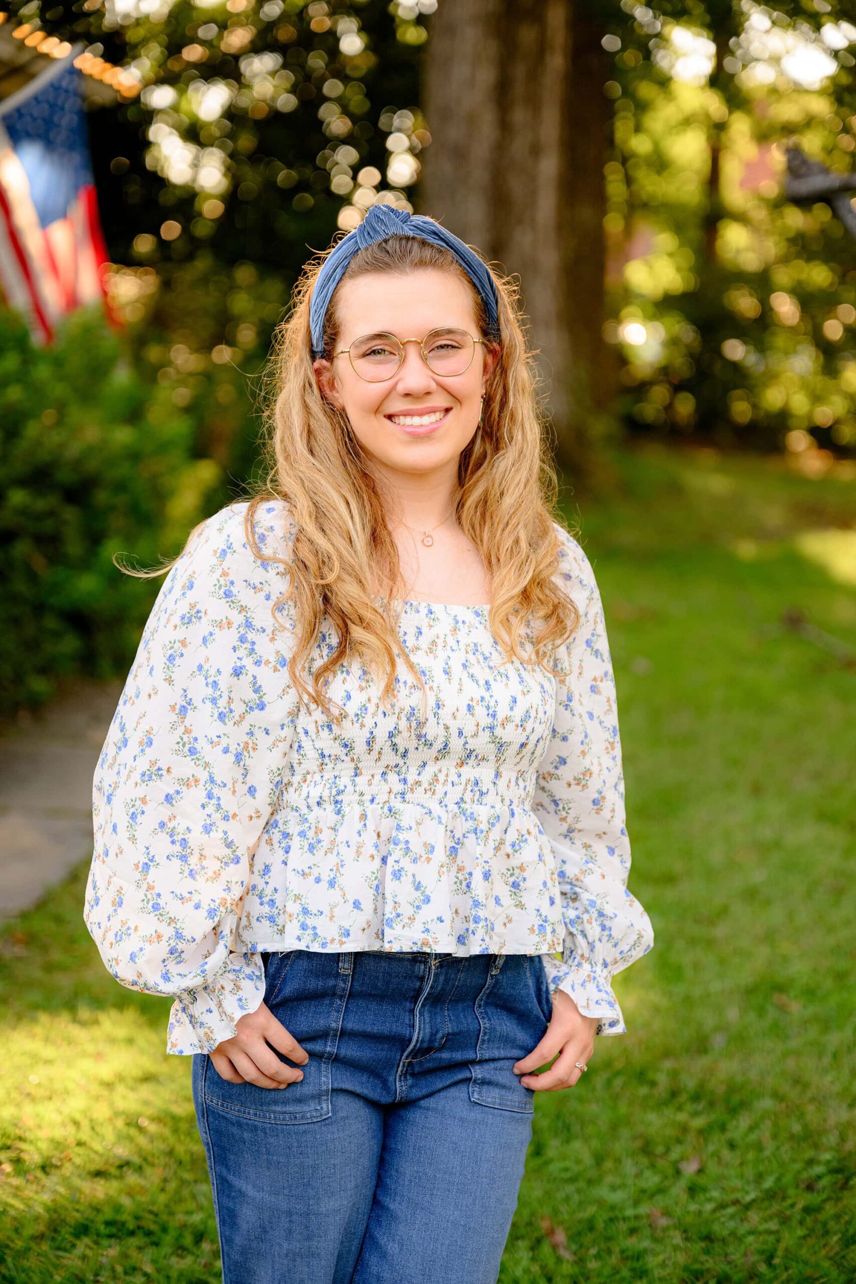 senior girl standing with her thumbs in her pockets and wearing a flower top and jeans with an american flag behind her blowing in the wind