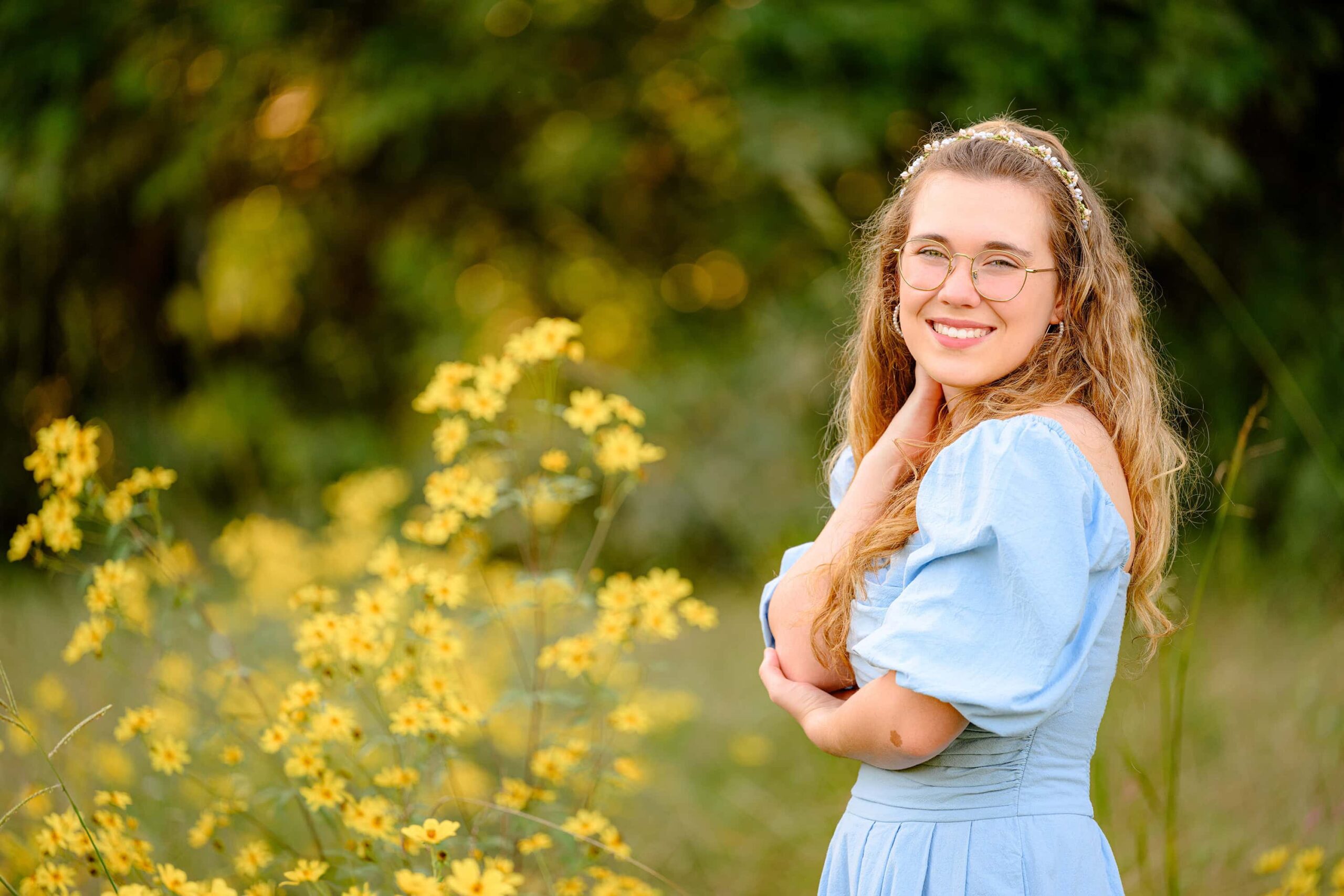 standing in a field of yellow flowers aubry looks over her shoulder smiling during her senior pictures wearing a blue dress and creative headband