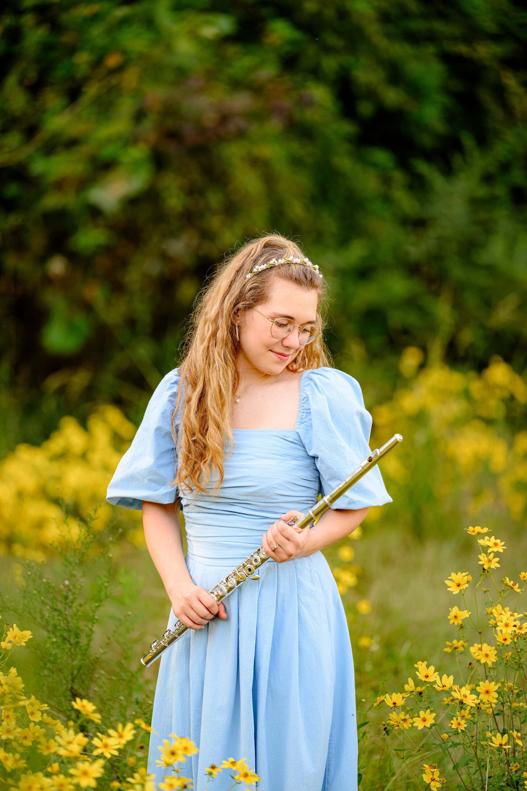 wearing a blue dress and beaded headband senior girl looks down at her flute, a meaningful prop she wanted to include in her grad session, while standing in a field with yellow flowers at a waxhaw park