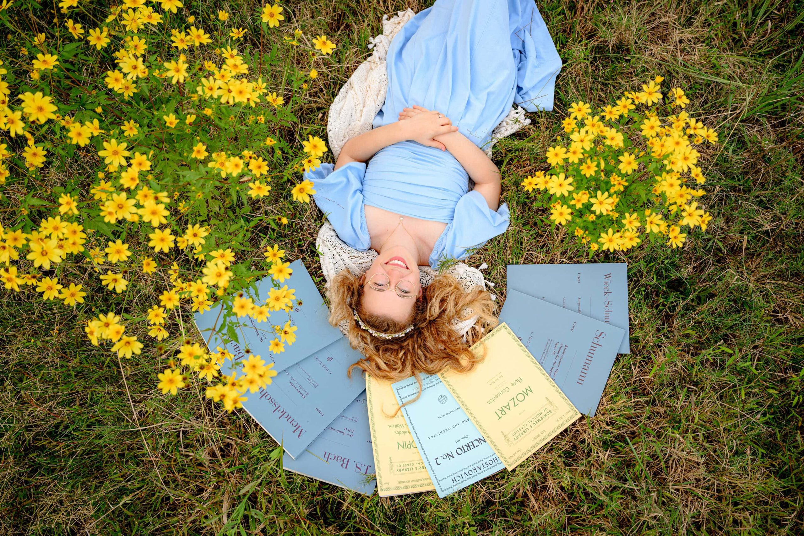 wearing a blue dress and surrounded by her favorite musical score books and yellow flower blooms lays in a field and smiles during her senior portrait session