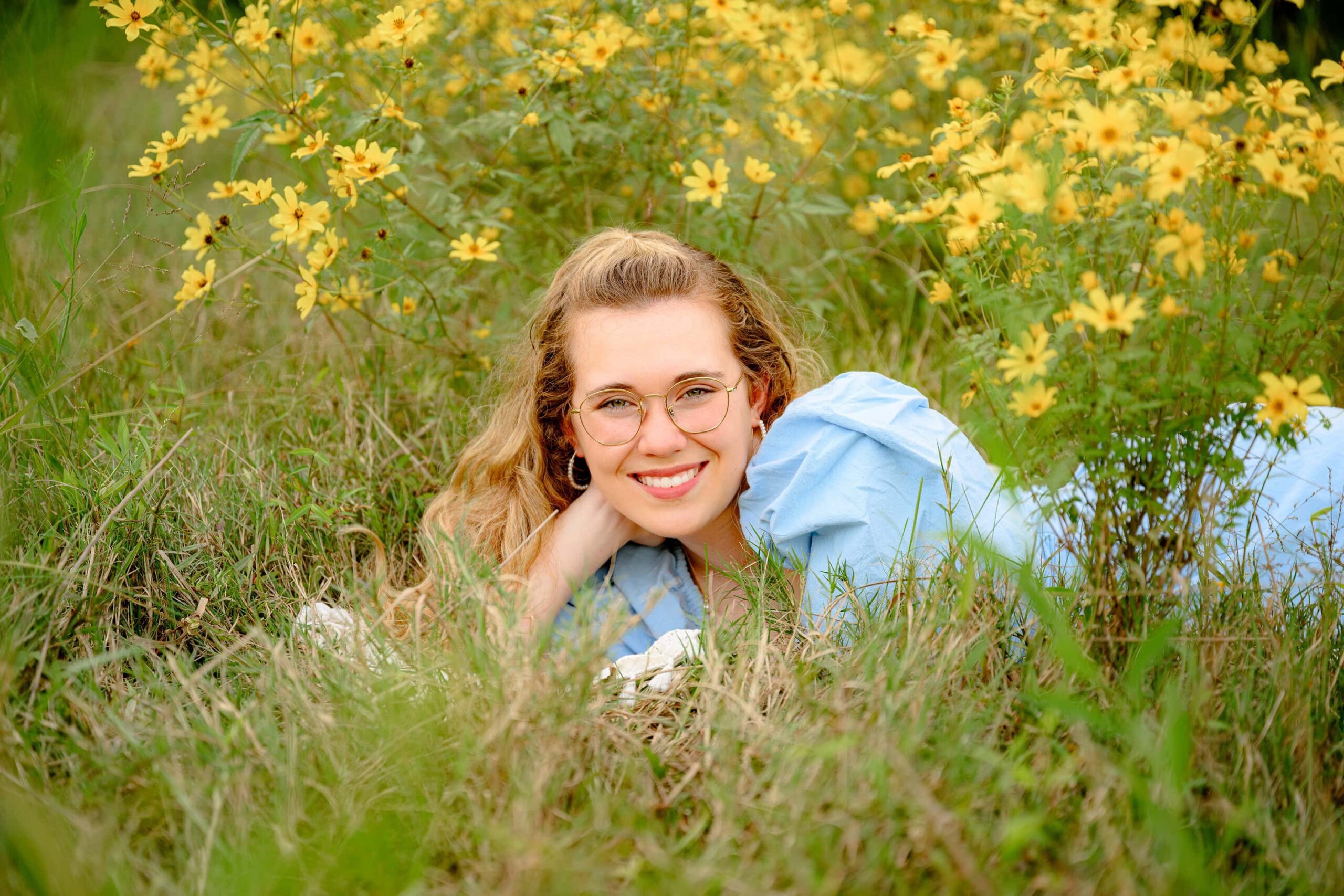 A grad poses creatively laying in a field flowing with yellow flowers wearing her glasses and blue dress