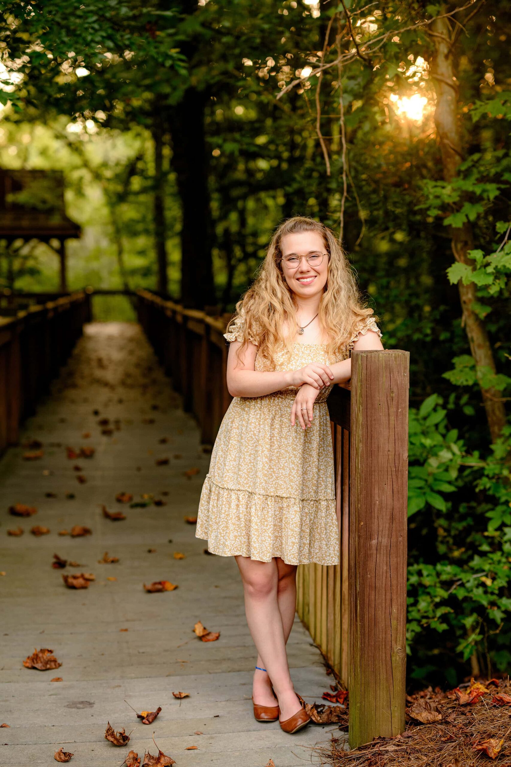 girl grad wearing a brown dress and shoes with her arms posed on the dark woodsy walkway during her senior portrait session