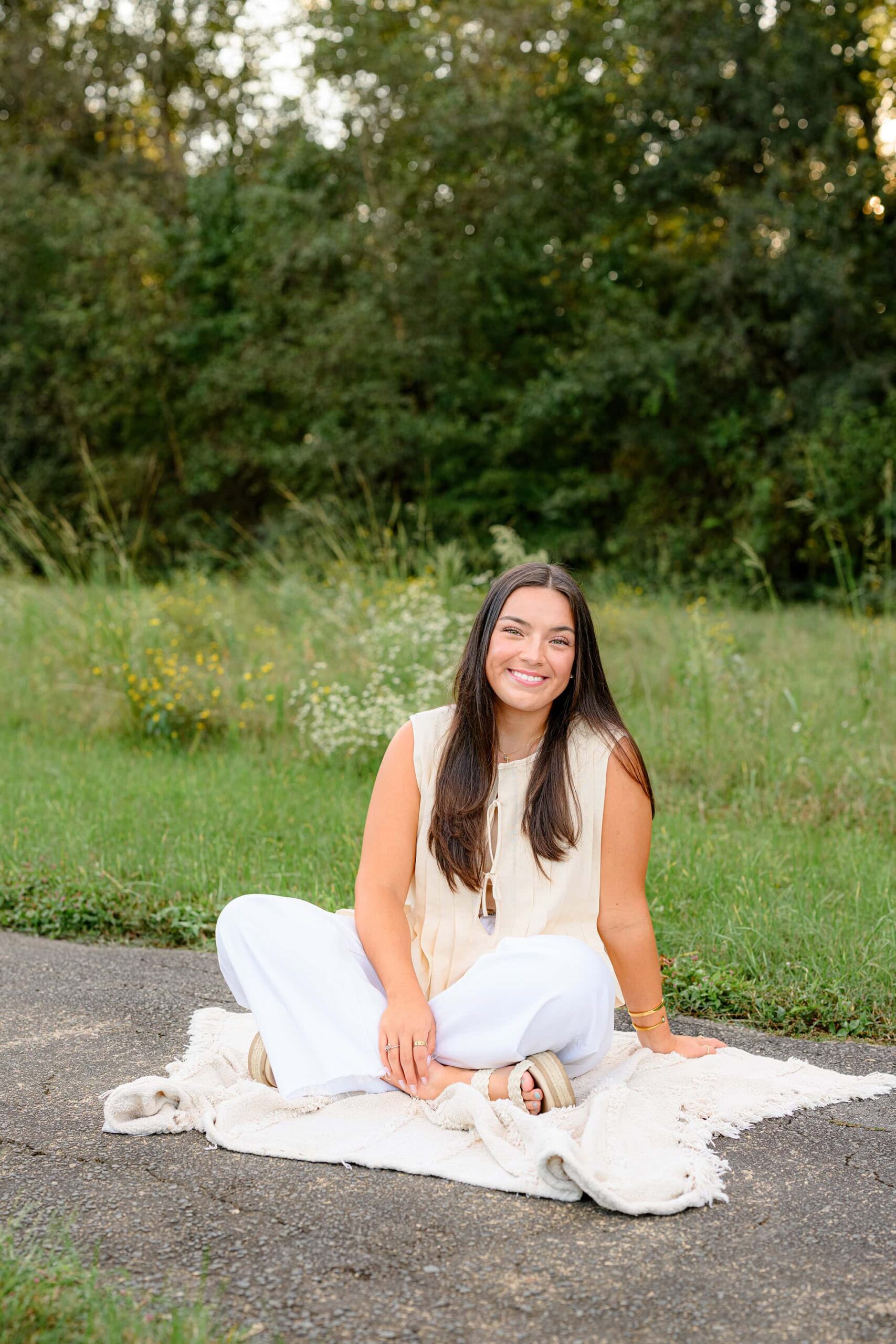 Senior photoshoot with a cuthbertson high school grad sitting on a blanket posing with one hand on the ground and the other in her lap wearing white pants and a cream shirt
