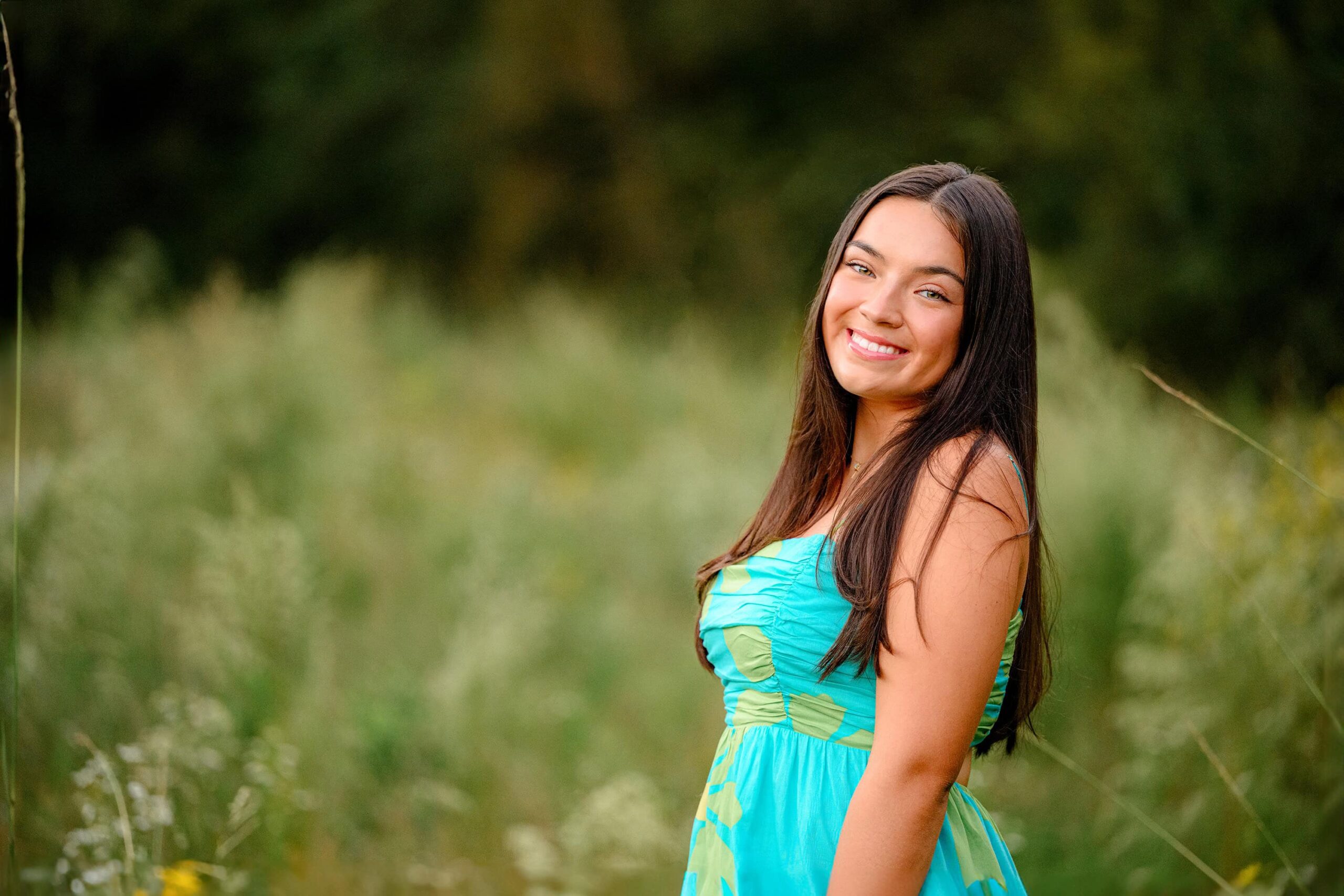 A Cuthbertson High School senior girl in teal dress looking over her shoulder with her hair hanging loosely while standing in a field of flowers poses for her grad photos