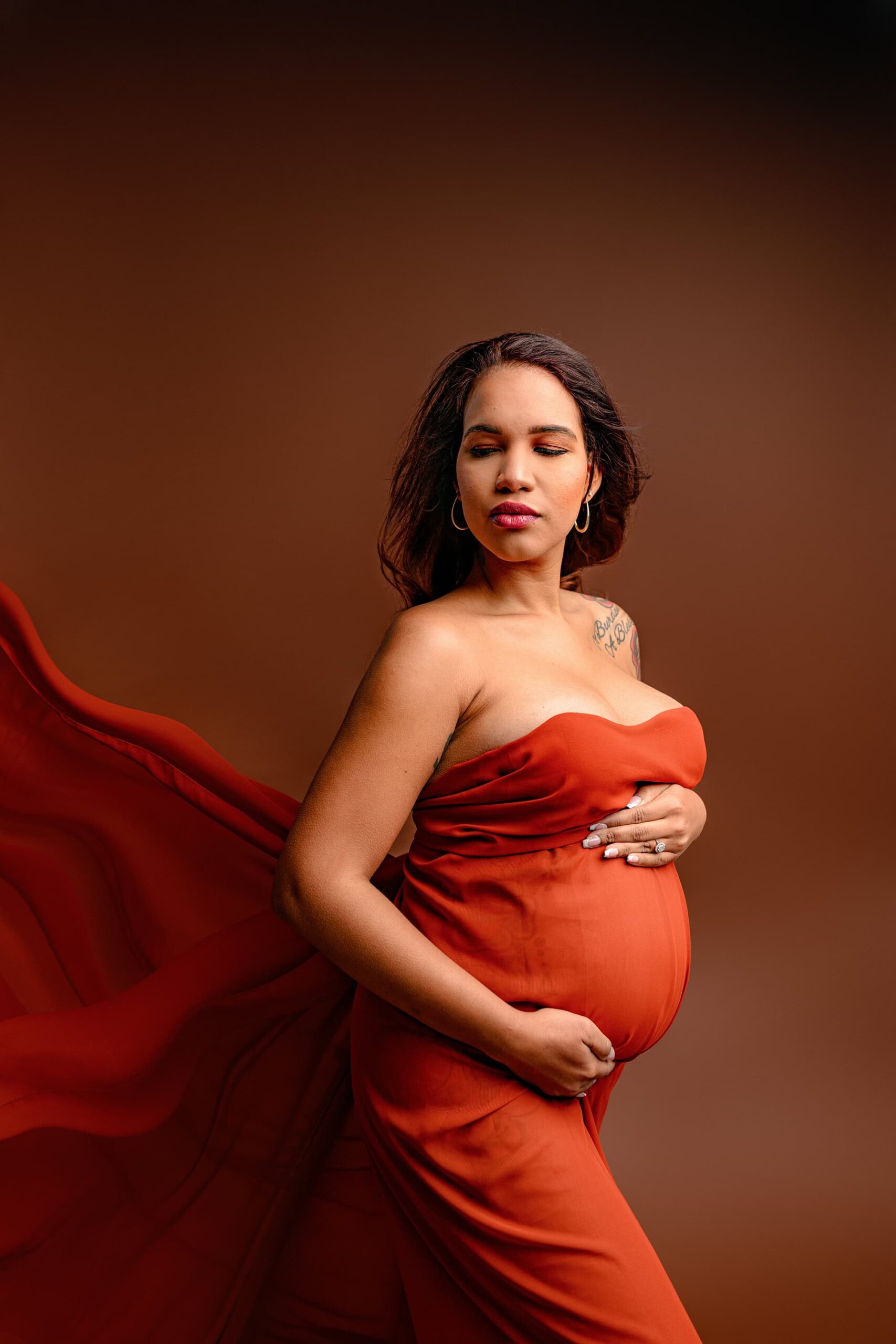 A captivating maternity portrait featuring the mama in a luxe, high-fashion pose. The rust chiffon flows behind her as she is photographed from the knee up in a semi side profile, highlighting her beautiful curves and radiant presence.