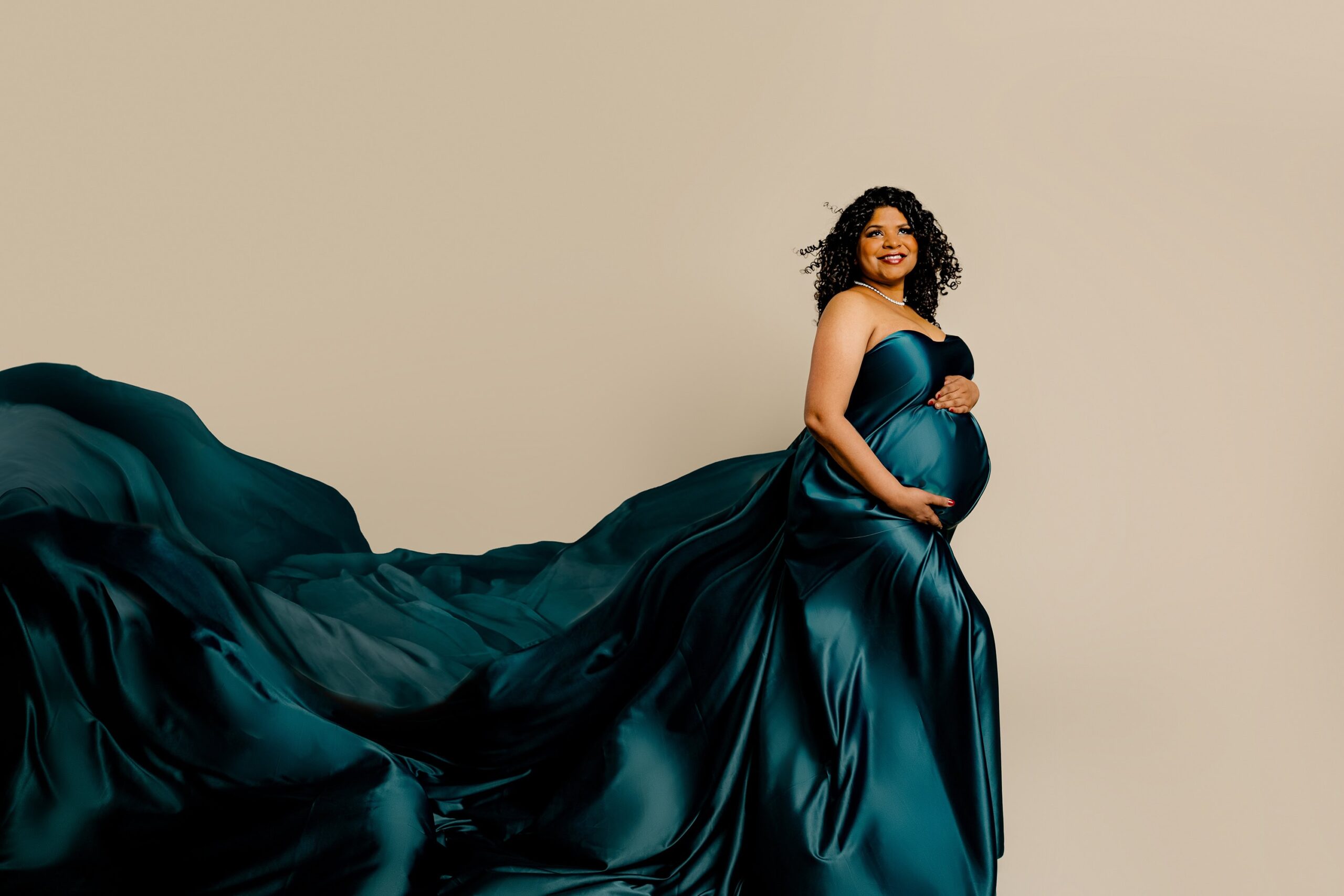 A radiant dark-haired mama poses in an elegant side profile against a soft cream background. Wrapped in flowing green silk, the fabric billows behind her, creating a sense of movement and timeless beauty while highlighting her glowing pregnancy.