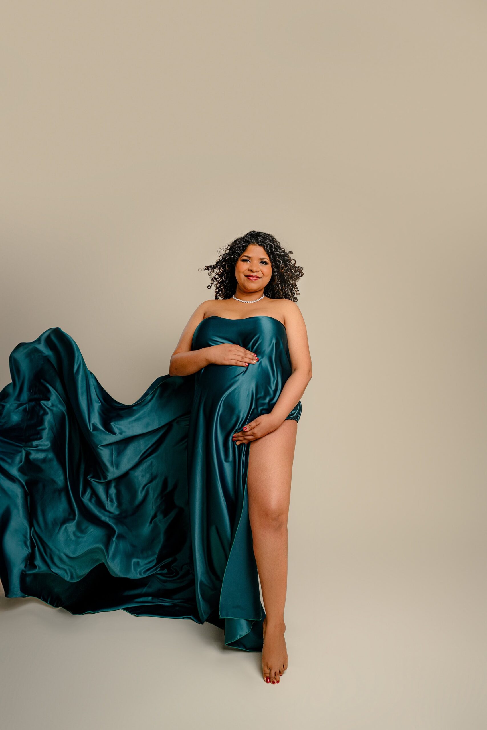 A stunning dark-haired mama poses against a soft cream background, wrapped in a rich green silk. With her belly beautifully accentuated and one leg gracefully exposed, the silk flows beside her, creating a timeless and sophisticated maternity image.