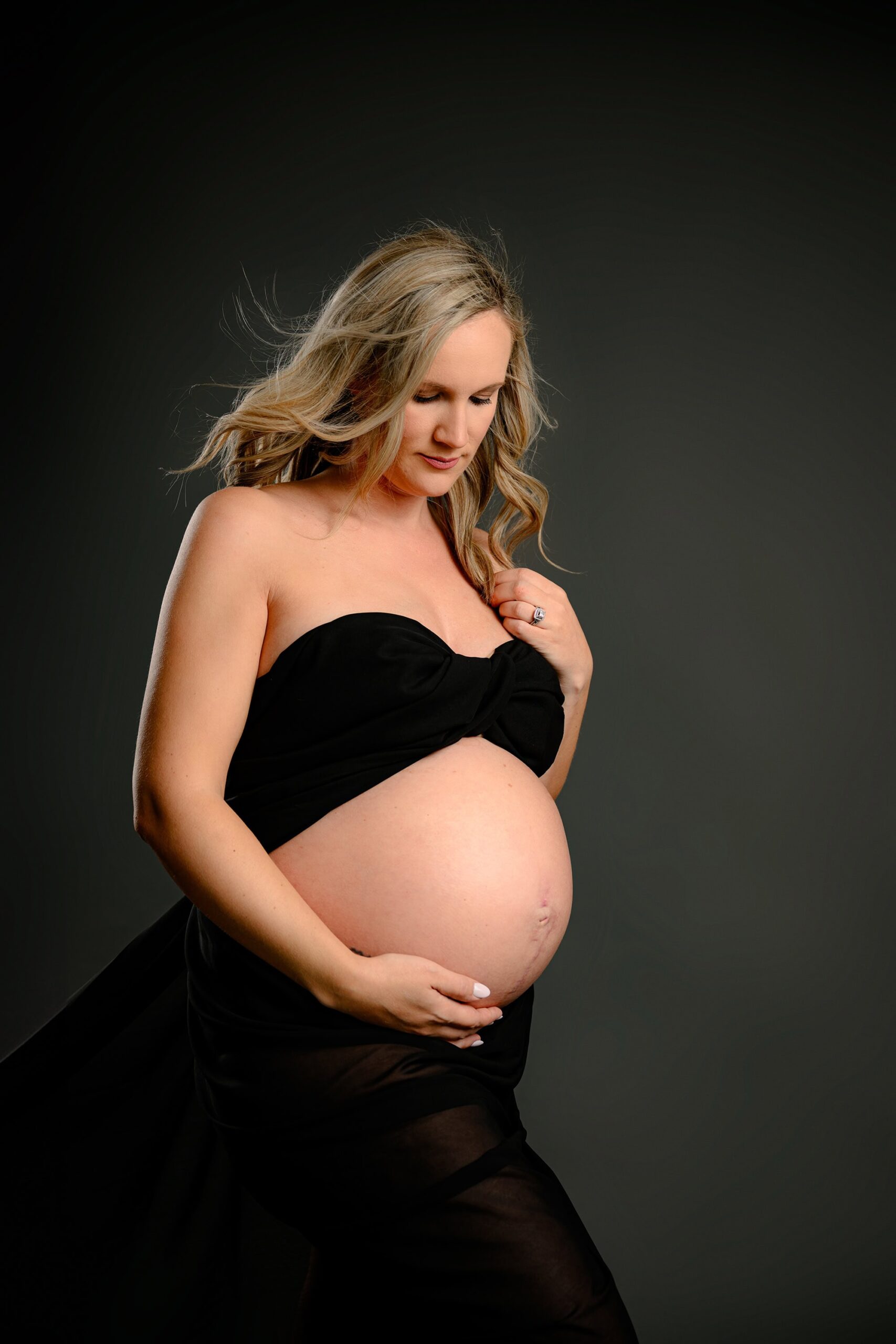 Looking down with a serene expression, this blonde mama gently places one hand in her hair and the other on the bottom of her belly. The flowing black chiffon adds a sense of movement, making this portrait both intimate and dramatic.