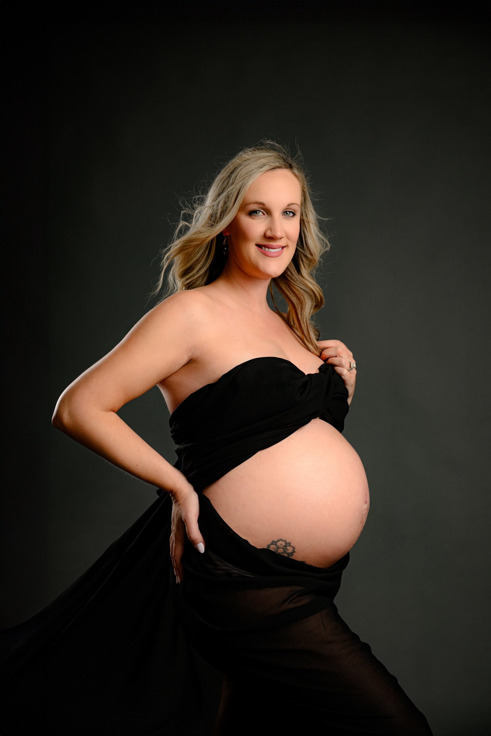 A stunning blonde mama poses against a moody grey backdrop, draped in sheer black chiffon. With one hand in her hair and the other resting on her hip, this striking image exudes confidence and elegance.