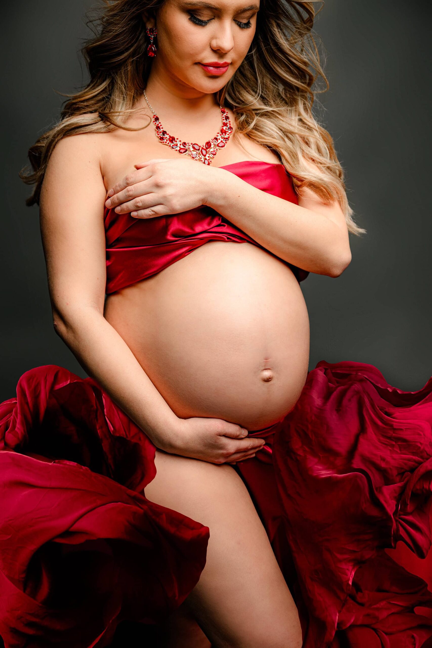 A stunning side-view shot focusing on her expectant belly wrapped in luxurious red silk. The soft draping of the fabric enhances the beauty of pregnancy, creating a striking and timeless maternity moment.
