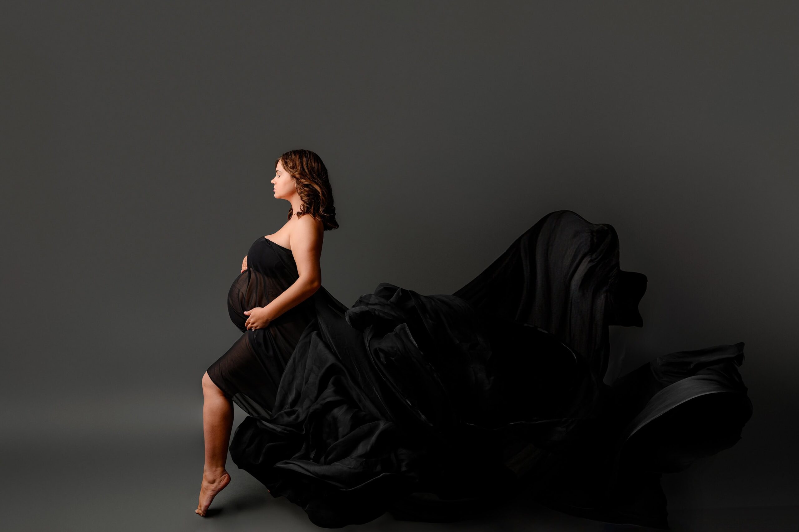 Full Body Profile Shot: A stunning maternity portrait of a mama with dark hair, wrapped in a flowing black chiffon wrap. She is turned to the side in a graceful full-body profile shot, with the chiffon trailing behind her, capturing a soft, elegant moment on a grey studio background
