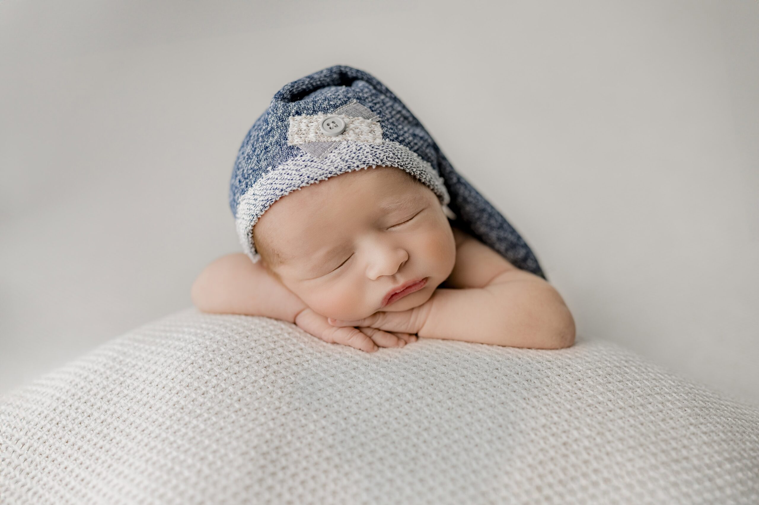 Newborn boy wearing a sleepy cap poses with his hands under his chin in waxhaw, nc studio