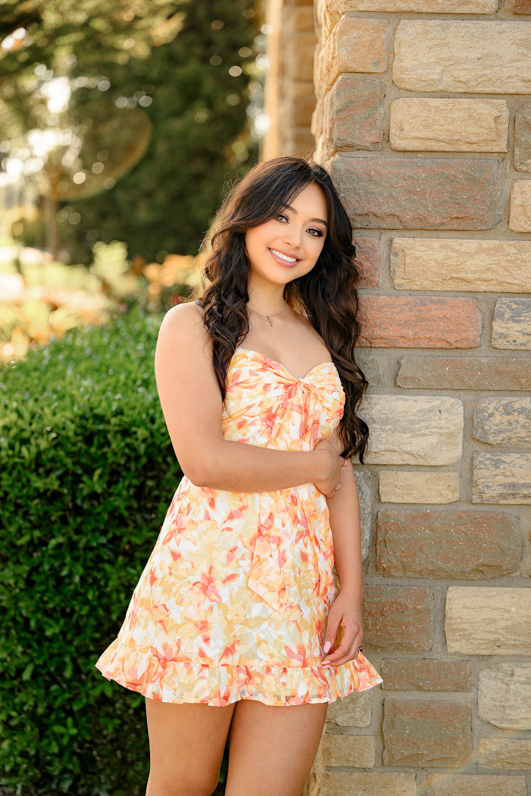 Senior girl stands with her hair, flowing down around her in a cute romper with orange and yellow toes and her hands wrapped around her waist