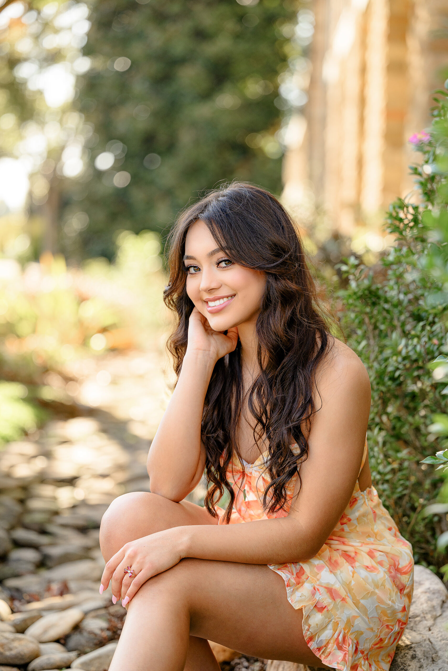 Senior girl sits looking towards the camera with her hand on her cheek, dressed in a orange and yellow romper