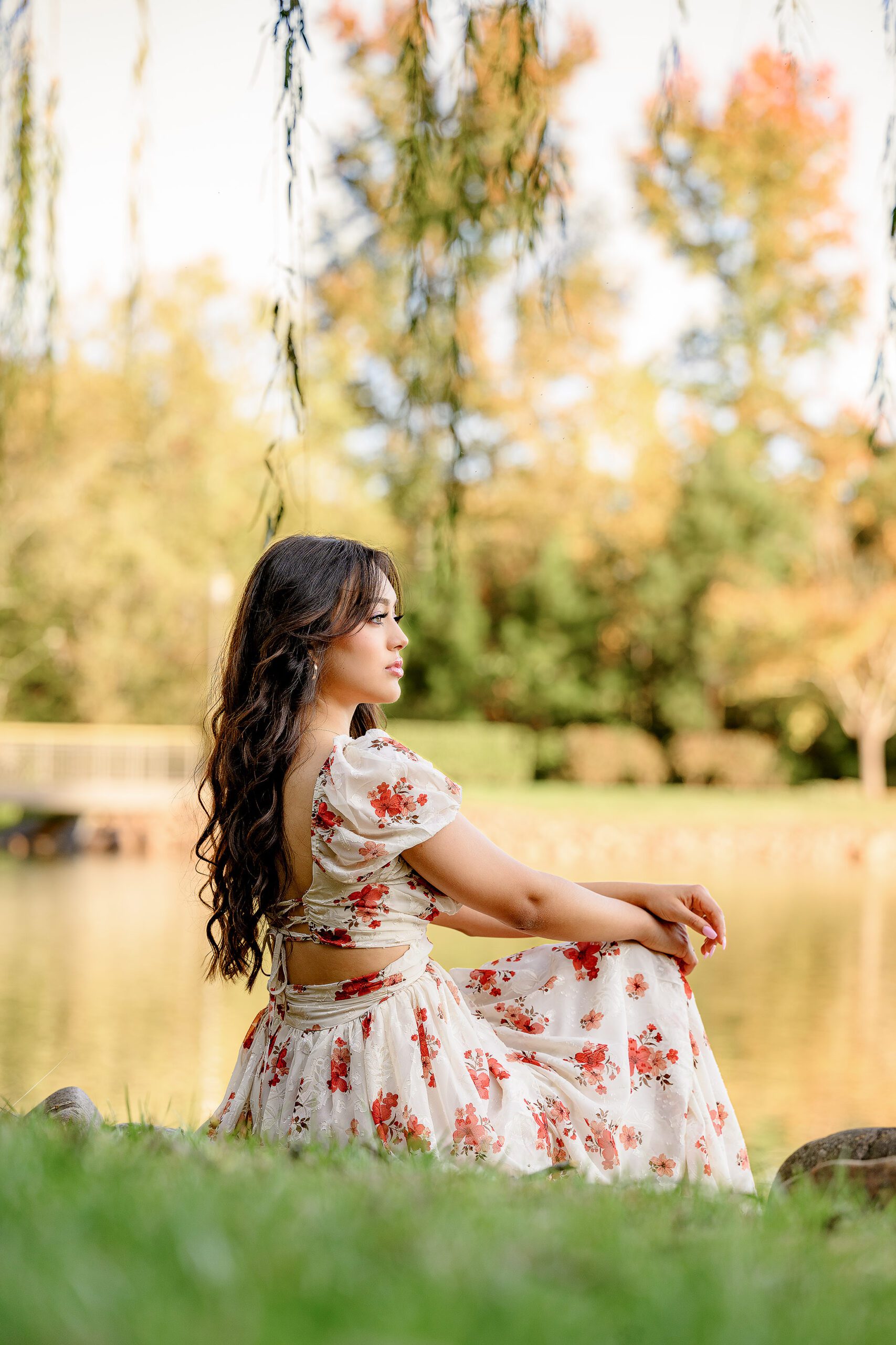 Senior girl sits on the grass with a pond and fall colors behind her long hair flowing behind her back, dressed in a white dress with red flowers